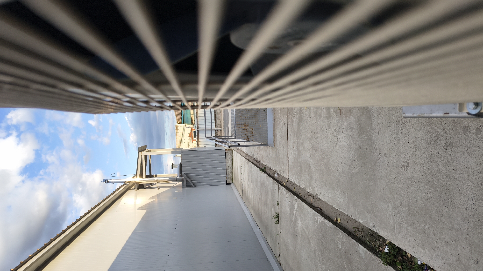 The image shows an outdoor scene with a long, grey, corrugated metal building on the left, casting a shadow on the adjacent concrete walkway. A metal pipe extends vertically from the roof of the building. To the right, a large, grey, slatted object, possibly an air conditioning unit, dominates the foreground, obscuring much of the view. A narrow concrete pathway runs alongside the building, with a trench or drainage channel running down its center, containing some sparse vegetation and debris. Further down the walkway, a metal ramp with handrails leads to a gate. In the background, a chain-link fence and some brick buildings are visible under a blue sky with scattered clouds. The location is identified as Mikhaylovka, Russia.