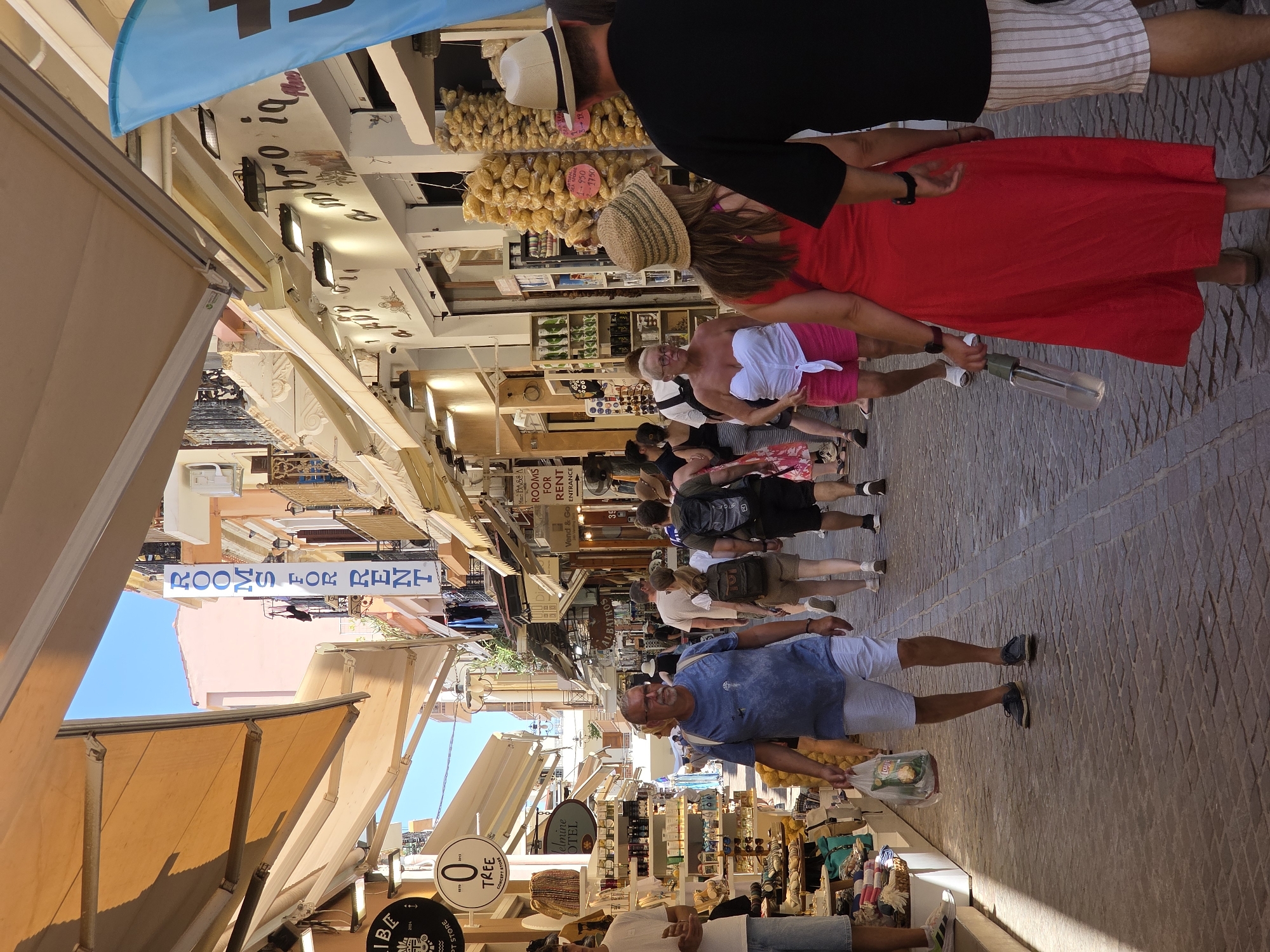 The image captures a bustling street scene in the city of Chania, Greece, likely within the historic Old Town or Venetian Harbor area, identifiable by its narrow cobblestone alleys, traditional architecture, and open-air shops. In the foreground, a man wearing a blue t-shirt and white shorts walks towards the viewer, carrying a plastic bag. To his right, a woman in a bright red dress and a straw hat walks away from the viewer, holding a clear bottle. Behind them, a man in a black t-shirt and striped shorts also walks away from the viewer. Several other people are visible walking along the street, browsing in shops, and moving in both directions, creating a sense of lively pedestrian activity. The street is lined with shops displaying goods such as souvenirs, food items (like dried fruits or nuts hanging from a display), and clothing. A prominent banner with "ROOMS FOR RENT" in blue lettering is visible hanging from one of the buildings. Awnings extend over the shops, providing shade from the sunlight, which casts shadows on the cobblestone pavement. The overall atmosphere is one of a vibrant tourist destination during daytime.