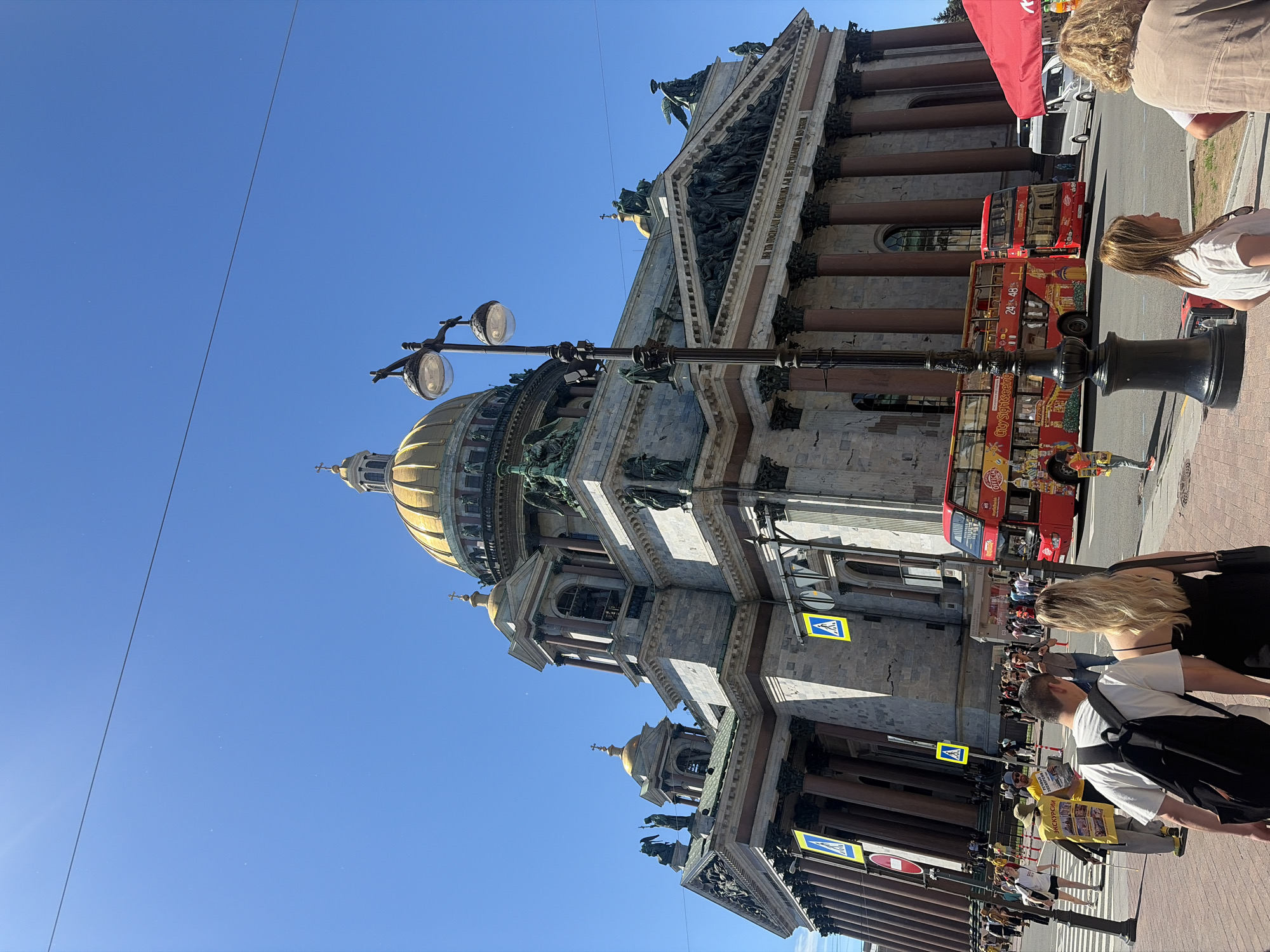 The image captures a wide-angle view of Saint Isaac's Cathedral in Saint Petersburg, Russia, under a clear blue sky. The imposing neoclassical cathedral, with its massive dome and intricate facade, is the central focus of the scene. In the foreground, a red double-decker tour bus is parked, its sides adorned with advertisements for "City Sightseeing." Several pedestrians are visible on the sidewalk and crossing the street. A lamp post stands prominently in the middle ground, and a red canopy is partially visible on the right side of the frame. The overall atmosphere is that of a busy tourist area on a sunny day.