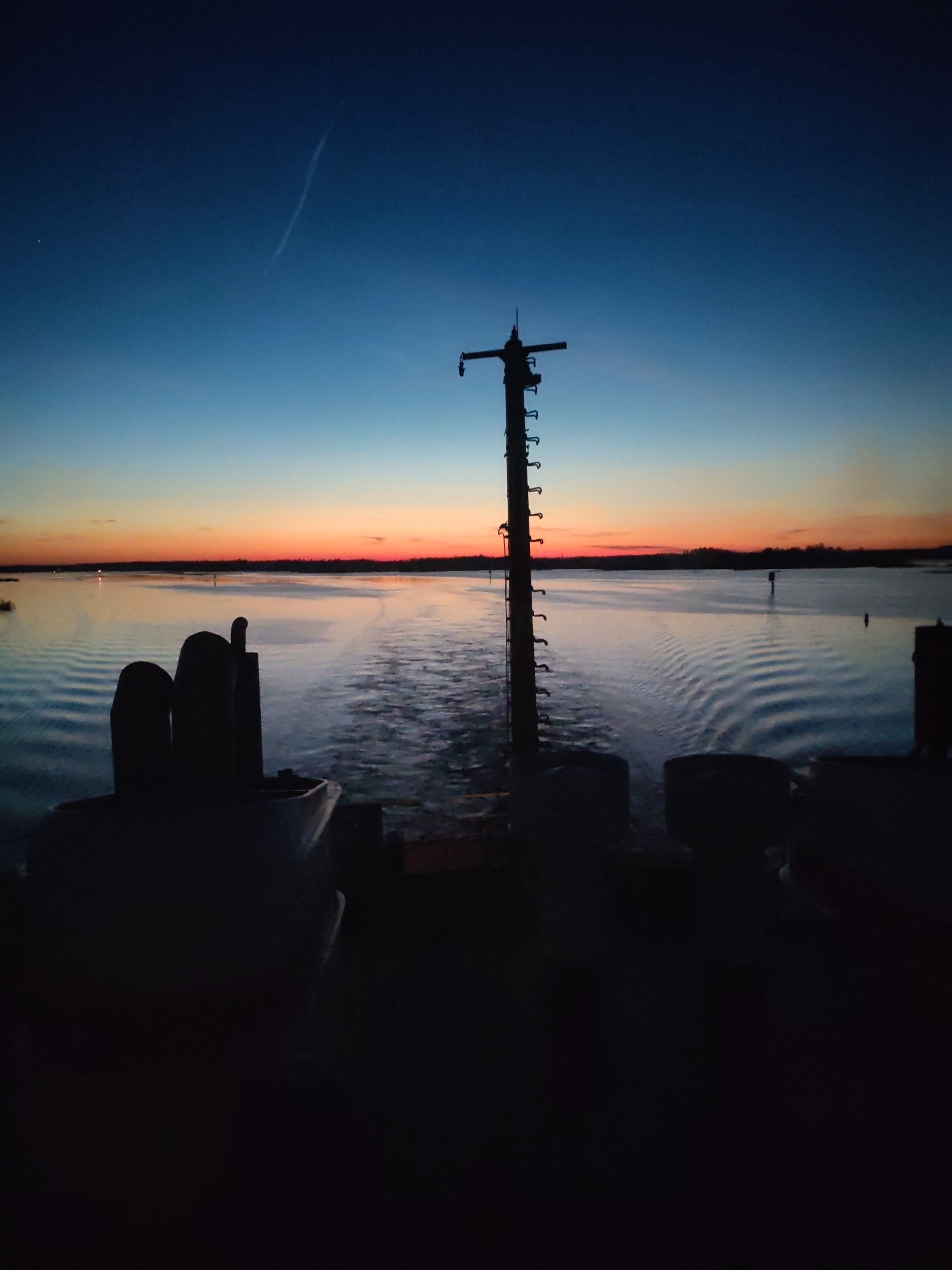 The photograph shows a view from a ship at sea, at dusk or dawn. The sky transitions from deep blue at the top to orange near the horizon. A condensation trail is visible in the upper center of the sky. The dark silhouette of the ship's mast and some other equipment is in the foreground. The water is calm, with visible wakes from the ship. There is a suggestion of land on the horizon. The specific location in Novokemskii, Russia, cannot be determined from the image.