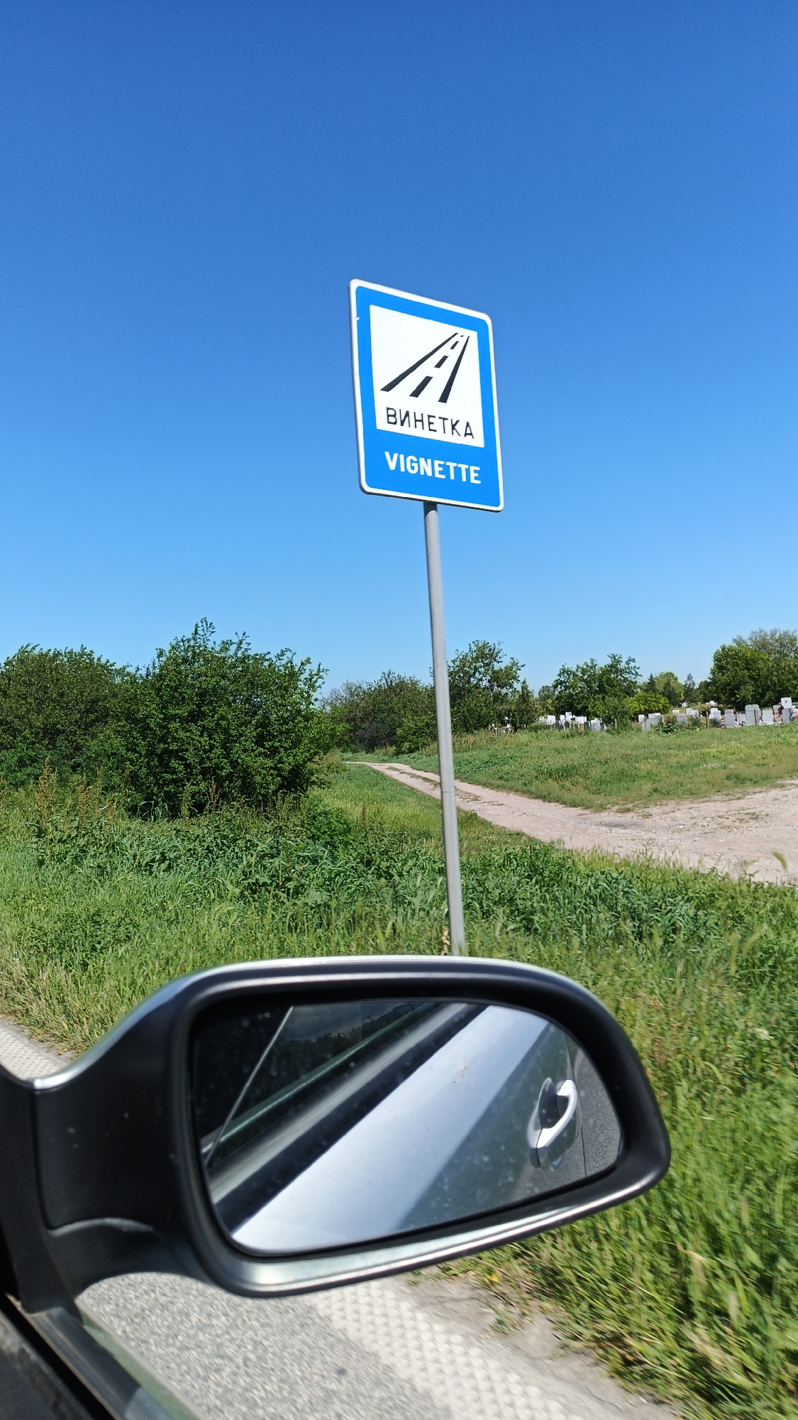 The image depicts a roadside scene, likely taken from inside a moving vehicle. A road sign indicating "Vignette" is prominently displayed against a clear blue sky. The sign features a stylized image of a highway. The foreground includes the side mirror of a car, partially obstructing the view. The background shows a grassy field with a dirt path leading towards a cemetery or similar area marked by rows of white or light-colored structures. Lush green vegetation is present on both sides of the road. The location is the city of Boliartsi, Bulgaria. No people are visible in the image.