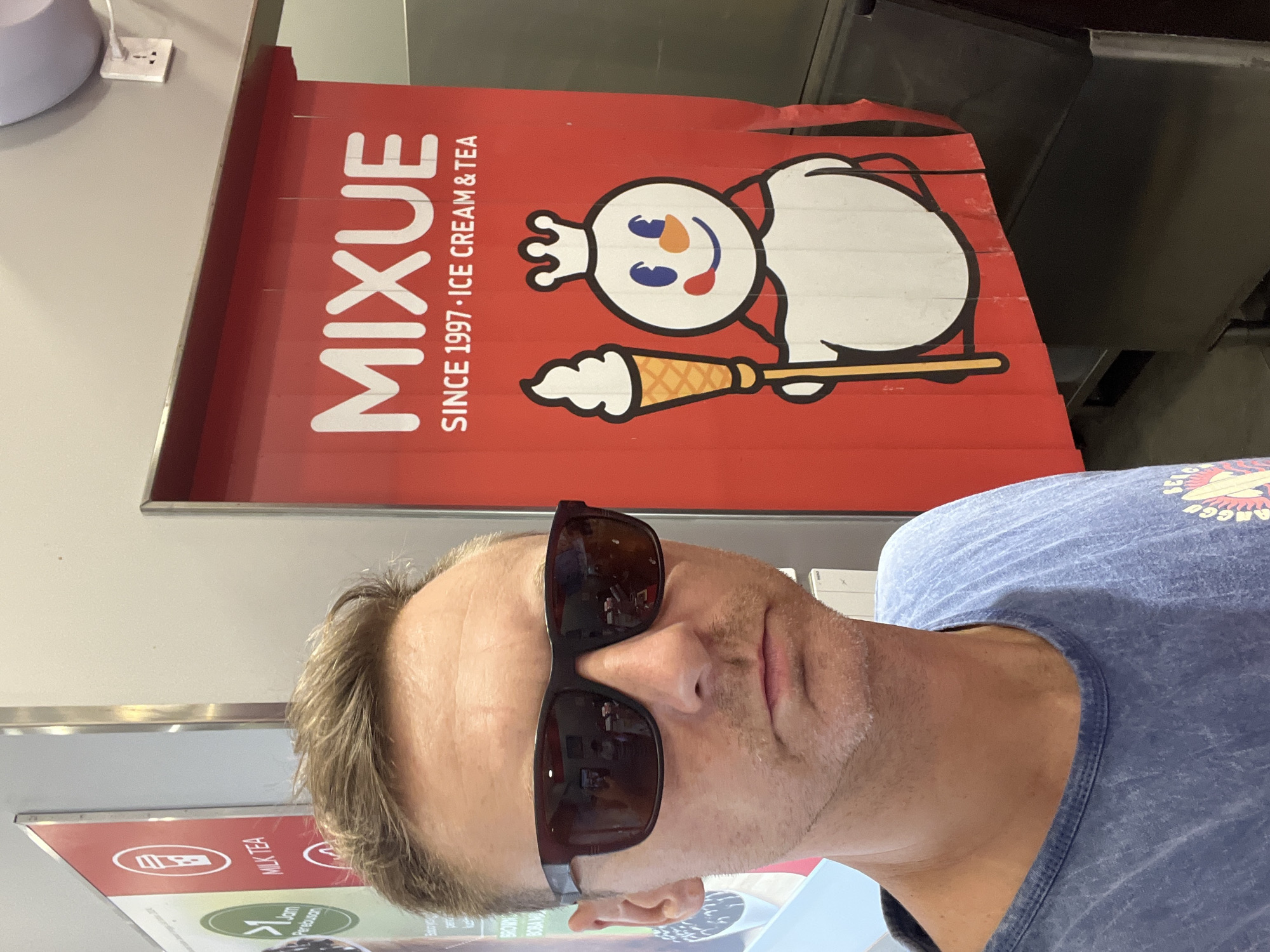 A man with fair skin and light brown hair, wearing sunglasses and a blue t-shirt, stands in front of a red banner advertising "MIXUE SINCE 1997 ICE CREAM & TEA". The banner features the brand's mascot, a snowman-like character wearing a crown and holding an ice cream cone. The background shows a store interior with other signage visible on the left, including the text "MILK TEA". The scene is likely in a commercial establishment in Sukawati, Indonesia, given the context. The man is the primary subject, positioned in the foreground. The lighting appears to be artificial, typical of an indoor setting.