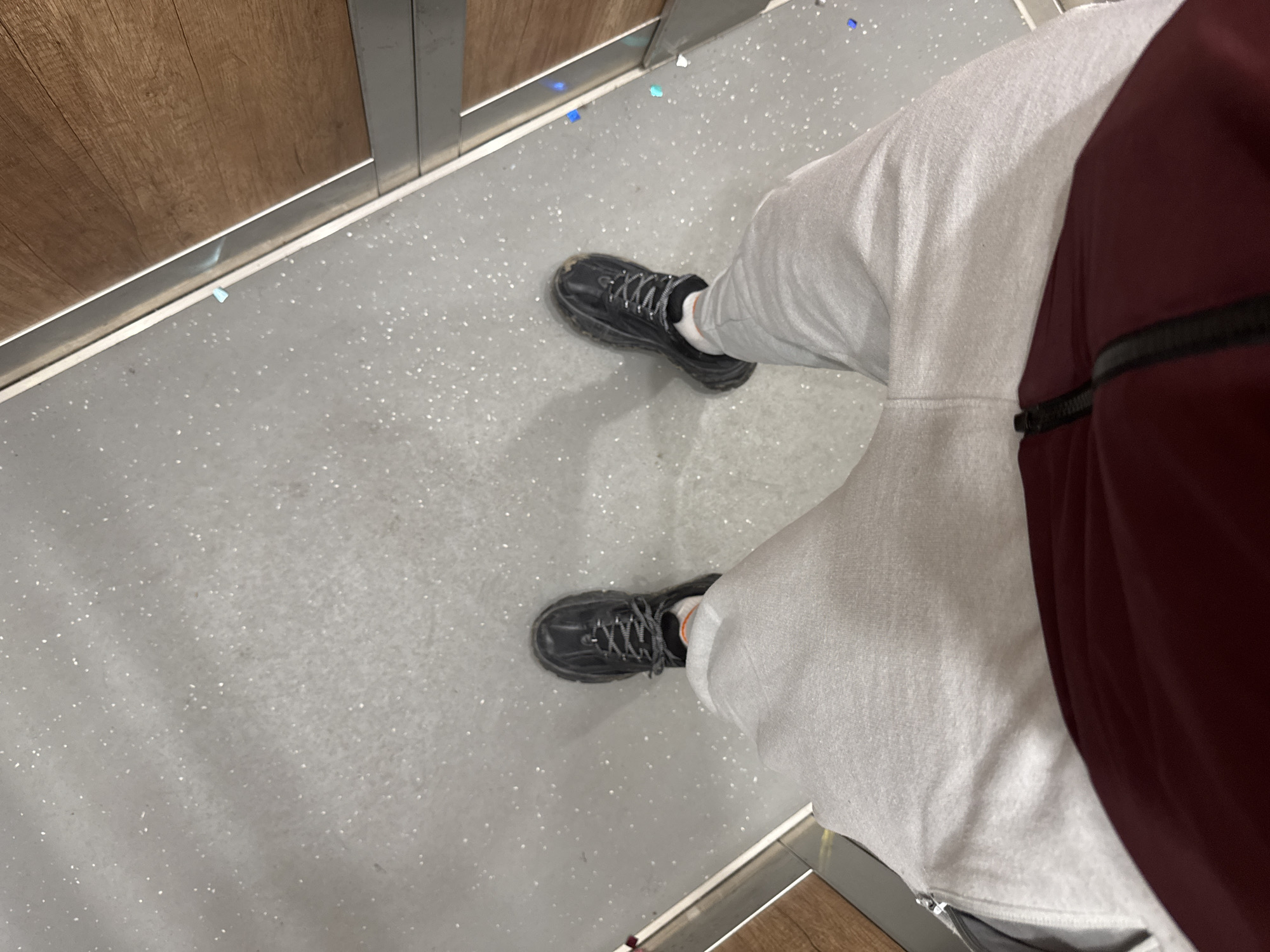 The image depicts a first-person perspective of a person standing indoors. They are wearing grey sweatpants, a burgundy jacket, and black sneakers with white laces and white socks. The floor is a light grey speckled surface, possibly linoleum or a similar material, covered in small glitter particles. The floor is bordered by sections of wood paneling. The location is unknown and cannot be determined from the image. The city of Sofia, Bulgaria is not relevant.