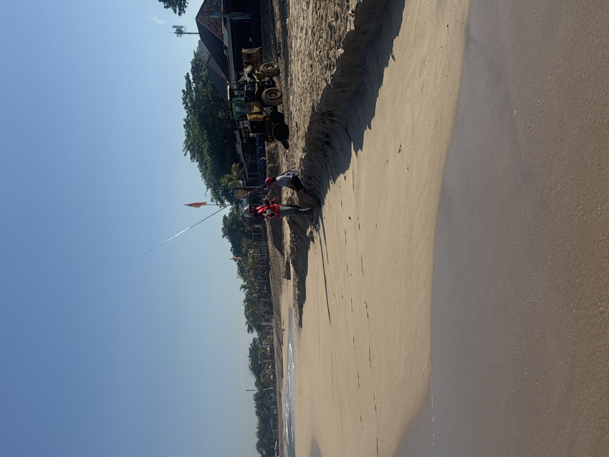 The image depicts a beach scene in Kuta, Indonesia. Two individuals, one wearing a red vest, appear to be working near a construction vehicle parked near a sand dune. An orange flag is planted nearby. The beach is wide with wet sand near the waterline. Trees line the back of the beach where buildings can be seen. The sky is clear.
