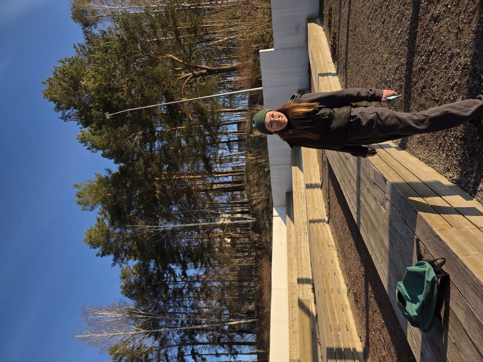 The image captures an outdoor scene featuring a young woman standing on a sunny day in Chaikovsky, Russia. The setting appears to be a park or a recreational area, characterized by a long wooden boardwalk or bench, alongside which runs a path of gravel and small stones.
The main subject is a young woman with long, reddish-brown hair, wearing a dark jacket, dark trousers, a dark green beanie hat, and glasses. She is smiling and looking directly at the camera, holding a light blue smartphone in her left hand. To her right, a dark green backpack rests on the wooden structure.
In the background, a dense forest dominates the landscape, composed primarily of tall coniferous trees, likely pines, with some bare deciduous trees intermingled. A clear, bright blue sky is visible above the tree line. A street lamp pole with a light fixture is also visible among the trees.
The long shadows cast by the person and the wooden structure indicate that it is either early morning or late afternoon, with the sun low in the sky. The overall atmosphere is bright and sunny, conveying a pleasant outdoor experience. No discernible text is visible in the image. The woman appears relaxed and happy, suggesting a casual outing.