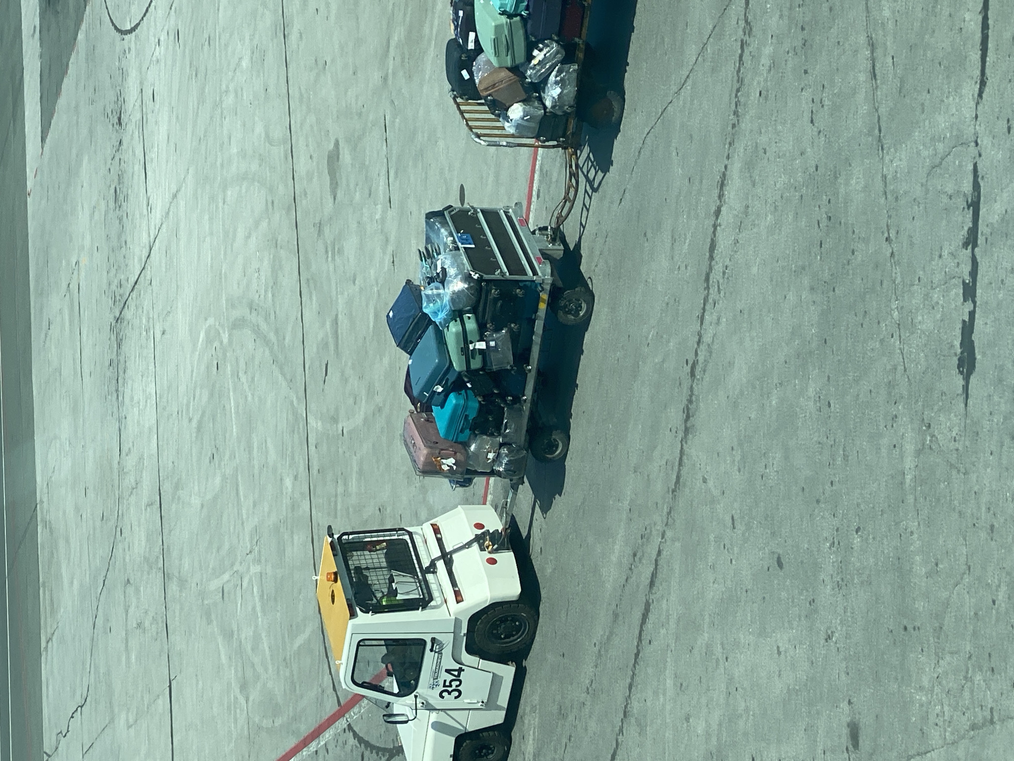 A white baggage tractor with the number 354 on its side tows two baggage carts loaded with luggage across a concrete tarmac. The first cart is a closed container filled with various suitcases, and the second cart is an open metal frame also laden with luggage. The scene is likely at an airport.