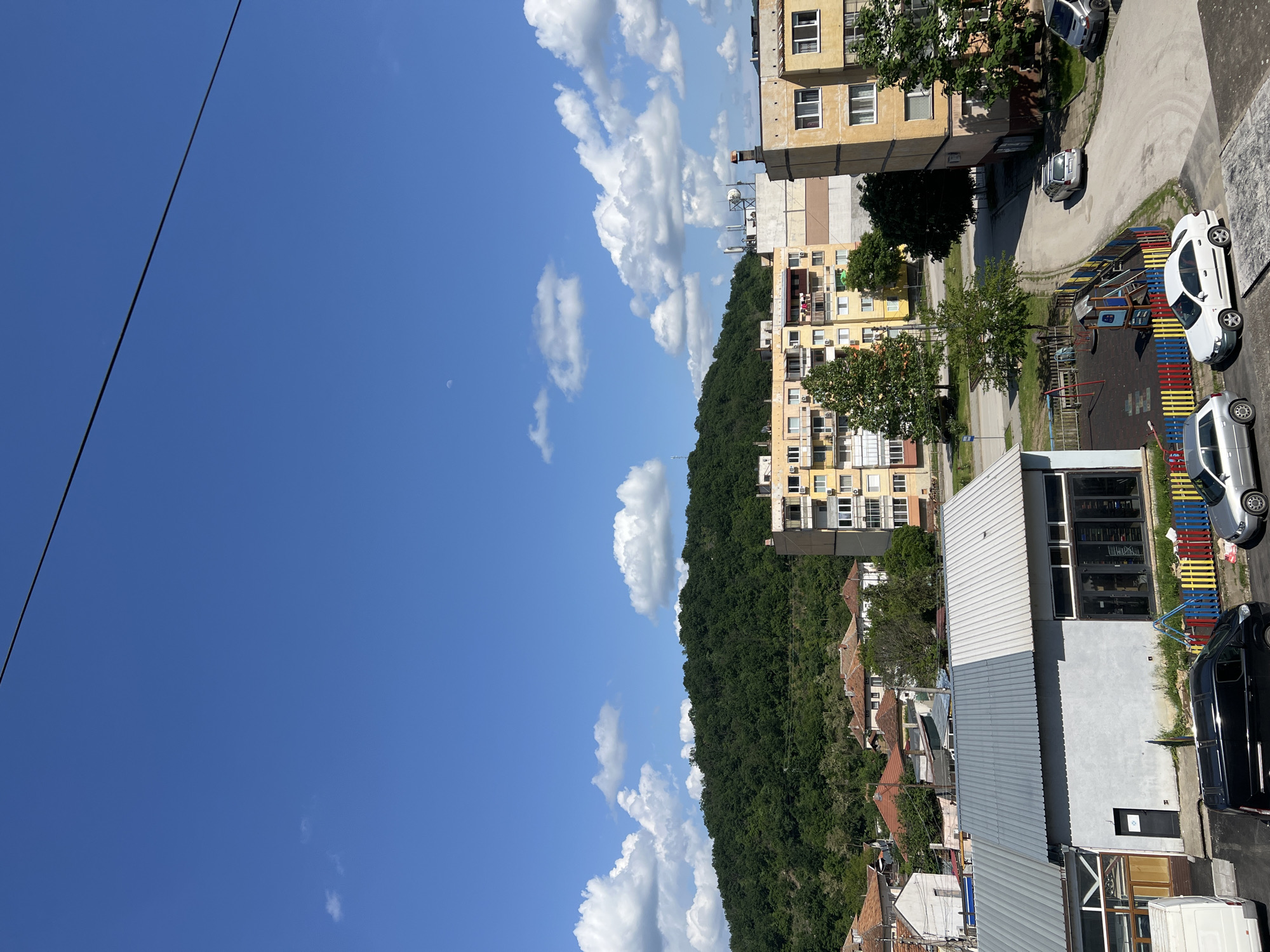 The image shows a daytime view of a neighborhood in the city of Roman, Bulgaria, observed from an elevated position. The sky is partially cloudy. On the right side of the frame are several apartment buildings and commercial structures. A wooded hillside is situated between the buildings and the sky. Cars are parked along the roadside and in a parking area. A playground with colorful barriers is visible adjacent to the buildings. The perspective is a mix of eye-level for the sky and birds-eye for the buildings and streets, indicating the shot was taken from a high vantage point like a rooftop or upper-story balcony. A powerline bisects the sky in the upper left area of the frame.