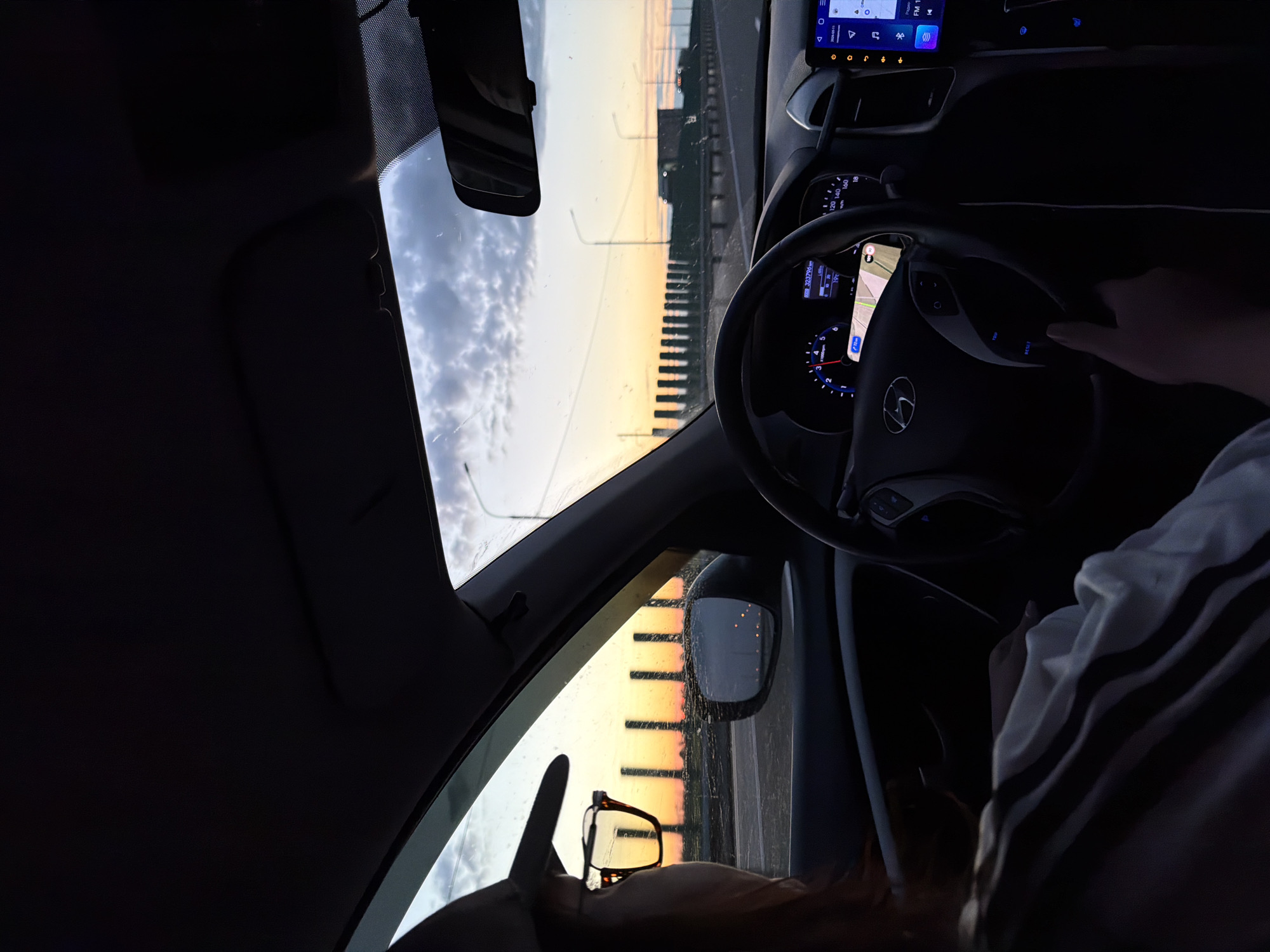 A view from inside a car driving along a road, potentially a bridge, at dusk or dawn. The driver is holding the steering wheel. The car's dashboard and infotainment system are visible. Through the windshield and side windows, the sky is lit with the colors of the sunrise or sunset, reflecting off the glass. A pair of eyeglasses rests on the dashboard in front of the passenger seat. Based on the map visible on the car's screen, the odometer reading, and other car details, it is difficult to pinpoint the exact location in Kislyakovskaya, Russia.