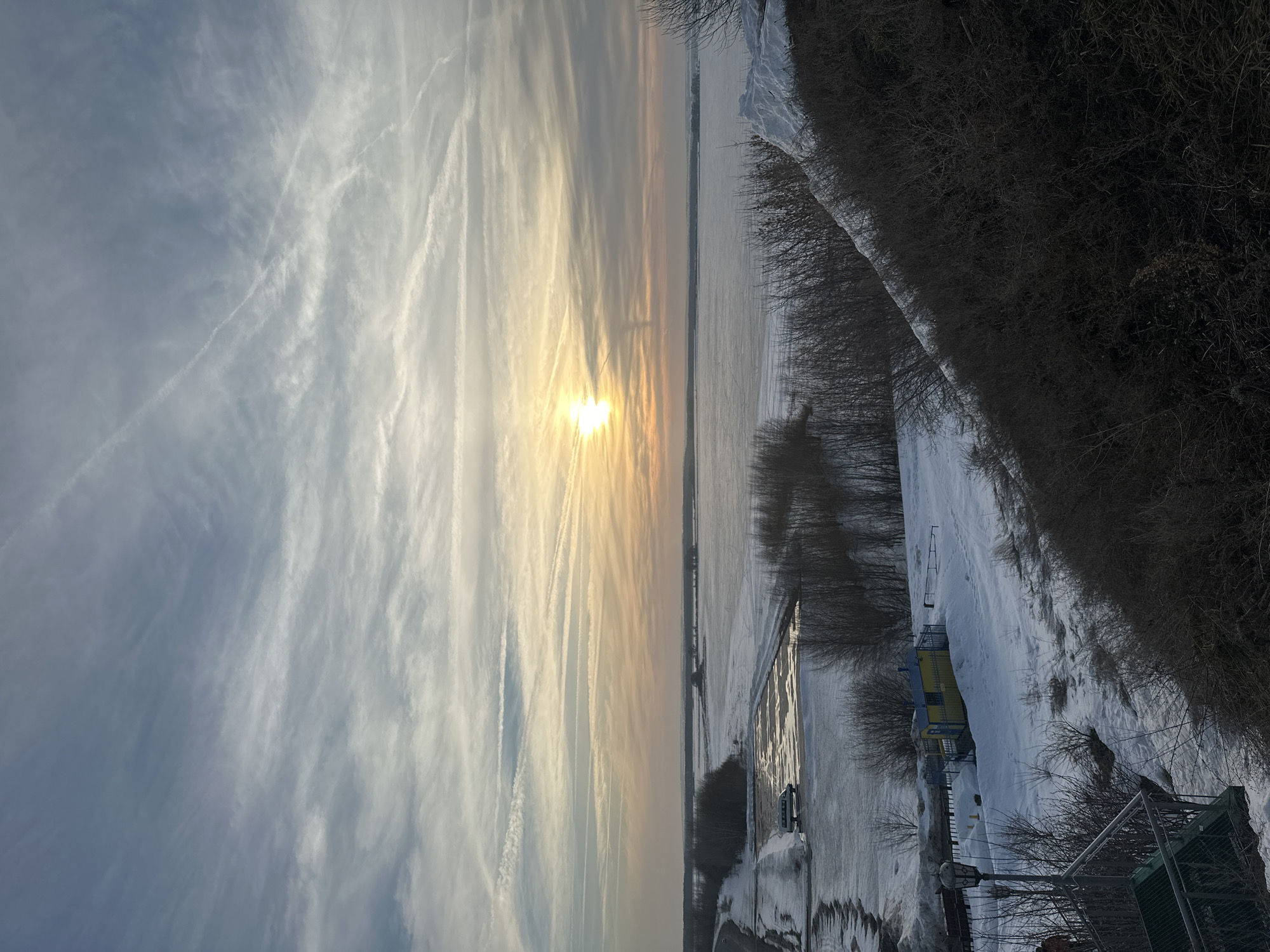The image depicts a panoramic winter landscape viewed from an elevated position in Sviyazhsk, Russia, at either sunrise or sunset. The sky, occupying the upper two-thirds, features a mix of pale blue, grey, and orange hues, with scattered cirrus clouds and multiple linear contrails. A bright, horizontally elongated sun, emitting a strong yellow-orange glow, is positioned slightly left of center on the horizon line.
Below the sky, a vast, mostly frozen body of water, likely the Sviyaga River or Kuibyshev Reservoir, stretches across the midground, covered in ice and snow. Patches of darker, possibly unfrozen water or slush are visible closer to the left bank. Distant land with a dark, bare tree line is discernible on the far horizon.
In the left foreground, a snow-dusted paved road or access area curves alongside the frozen expanse. A light-colored van or small bus is parked on a partially melted section of this pavement. To the immediate right, a steep, snow-covered embankment descends, densely populated with dark, bare deciduous shrubs. A significant pile of white snow rests atop this embankment in the upper right quadrant.
The lower left foreground reveals a metal staircase with green grate steps and handrails leading downwards. Adjacent to the staircase, a black, ornate streetlamp stands upright. Further down the slope, below the road level, a small, yellow utility building with a blue roof and blue trim is enclosed by a dark metal fence. The ground in the foreground is covered in snow, showing visible tracks. No individuals are present in the scene.