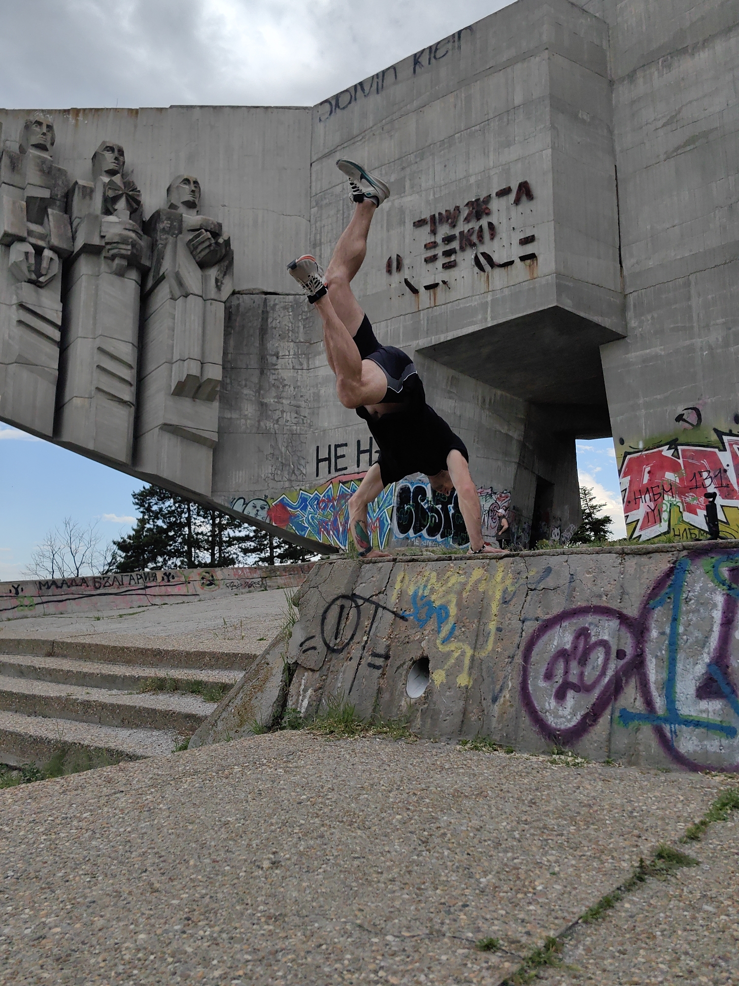 A man is performing a handstand on a graffiti-covered concrete ledge. The ledge is part of a larger, dilapidated concrete structure. The structure includes a relief of several human figures. Graffiti covers much of the surface of the structure. There is a staircase leading down from the ledge. Trees and a cloudy sky are visible in the background. The location is the Pantheon of the Bulgarian Revival Heroes in Varna, Bulgaria.