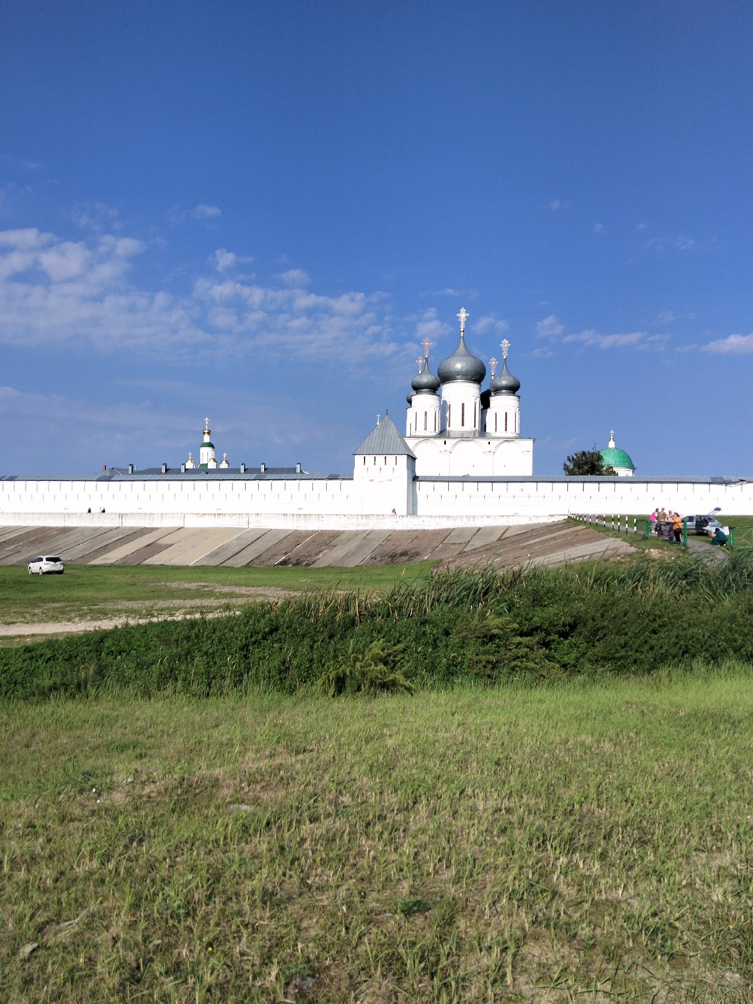 The image depicts the Makar'evo Monastery in Makar'evo, Russia, under a clear blue sky with scattered white clouds. The monastery features a prominent white fortress wall, beyond which a group of white buildings with gray domes topped with crosses are visible. One building has a distinct green dome. In the foreground, a grassy field slopes down towards the viewer, with a path and a white car on the left side. On the right side of the fortress wall, a group of people are gathered near a fence and vehicles. The overall scene suggests a sunny day at a historical and religious site.