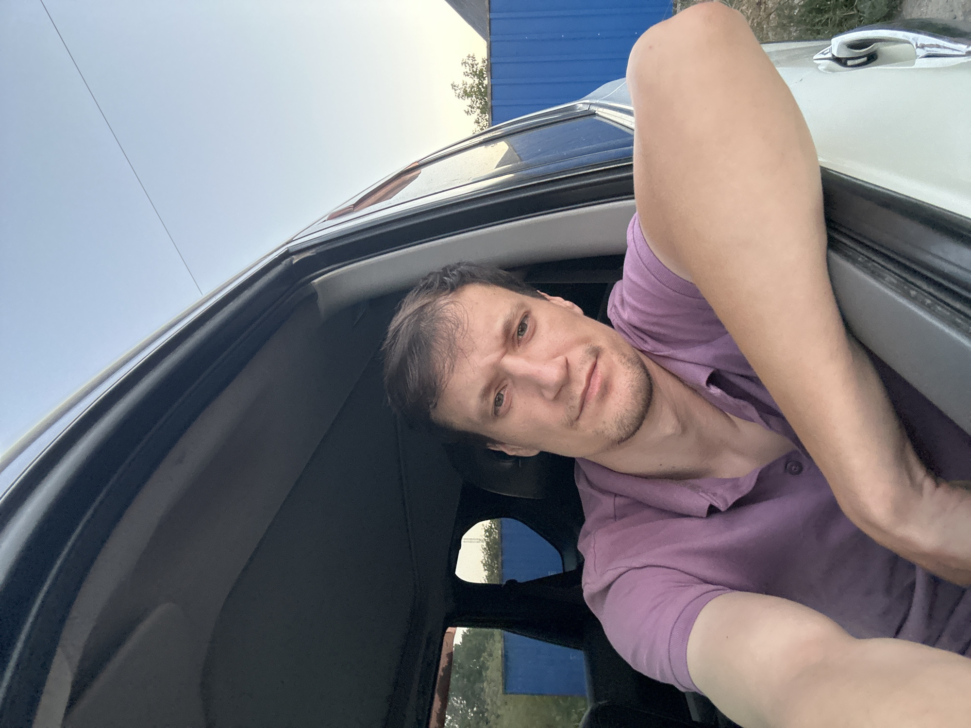 A man wearing a purple polo shirt is sitting in a car, with his right arm resting on the open car door. He is looking towards the camera with a neutral expression. The car is parked outdoors, with a blue fence and some trees visible in the background. The sky is clear and blue. The image appears to be a selfie taken in the city of Shatava, Ukraine.