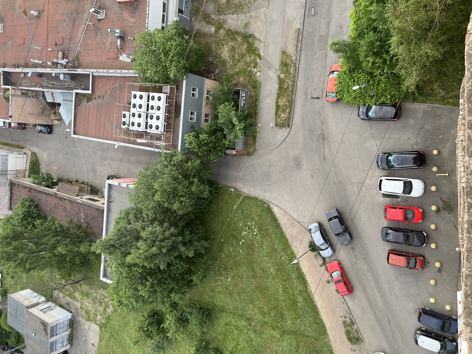 This is a high-angle, daytime aerial view of an urban setting in Sinyavino, Russia. The scene is dominated by a mix of residential buildings, industrial structures, and abundant greenery. Several cars are parked on paved areas surrounding the buildings, including a red hatchback, a black sedan, a dark blue sedan, and a white sedan. One building has a red-tiled roof with numerous outdoor air conditioning units. Another building is a smaller, light blue structure with windows, partially obscured by trees. A patch of grassy area is visible, with some scattered debris. Overhead power lines crisscross the scene. The lighting suggests it is daylight, likely overcast or with diffused sunlight. No people are visible, and the overall impression is one of a quiet, possibly residential or mixed-use area.