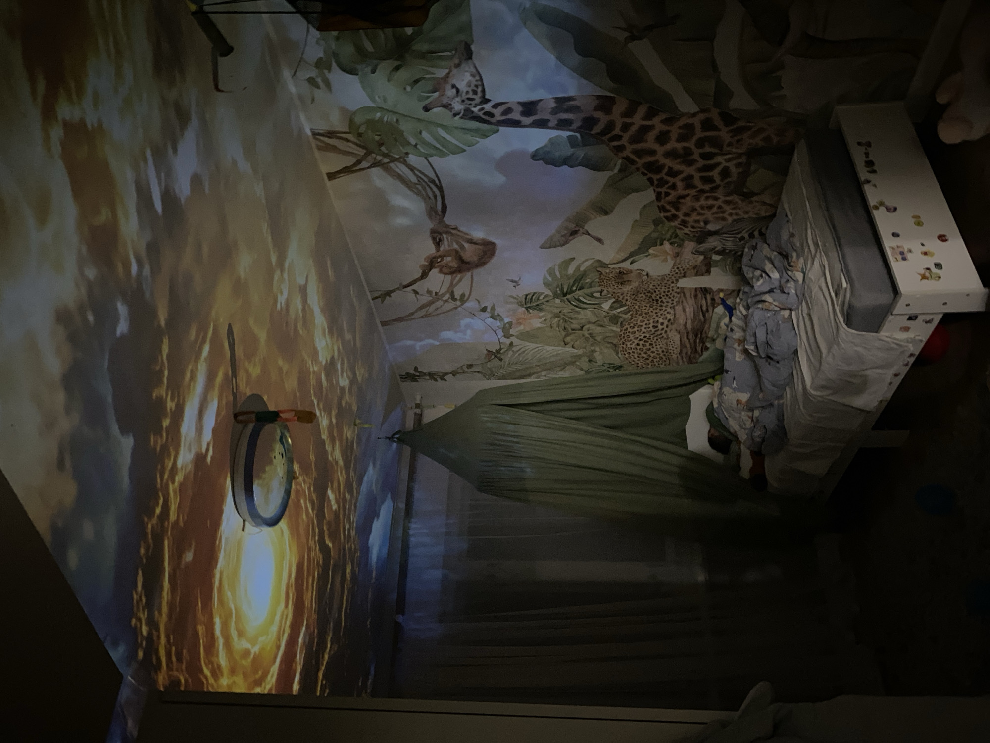The image displays a dimly lit child's bedroom in Chaikovsky, Russia. The ceiling features a projected scene of a cloudy sky with prominent orange and yellow hues, resembling a sunset or sunrise. A circular light fixture or projector is mounted on the ceiling, actively casting this image. The right wall is adorned with an extensive jungle-themed mural or decals, depicting a spotted leopard positioned lower left, a tall giraffe on the lower right, and a sloth hanging from vines in the upper middle, all surrounded by large green leaves and other foliage. A white-framed child's bed is situated against this decorated wall, where a child is observed sleeping under a blue and white patterned duvet, partially covered by an olive-green conical fabric canopy suspended from the ceiling. The bed's lower frame includes a pull-out drawer embellished with multiple colorful stickers. On the left side of the room, a sheer, dark curtain covers a window or opening. The floor is dark, with a faint blue object visible in the lower left corner.