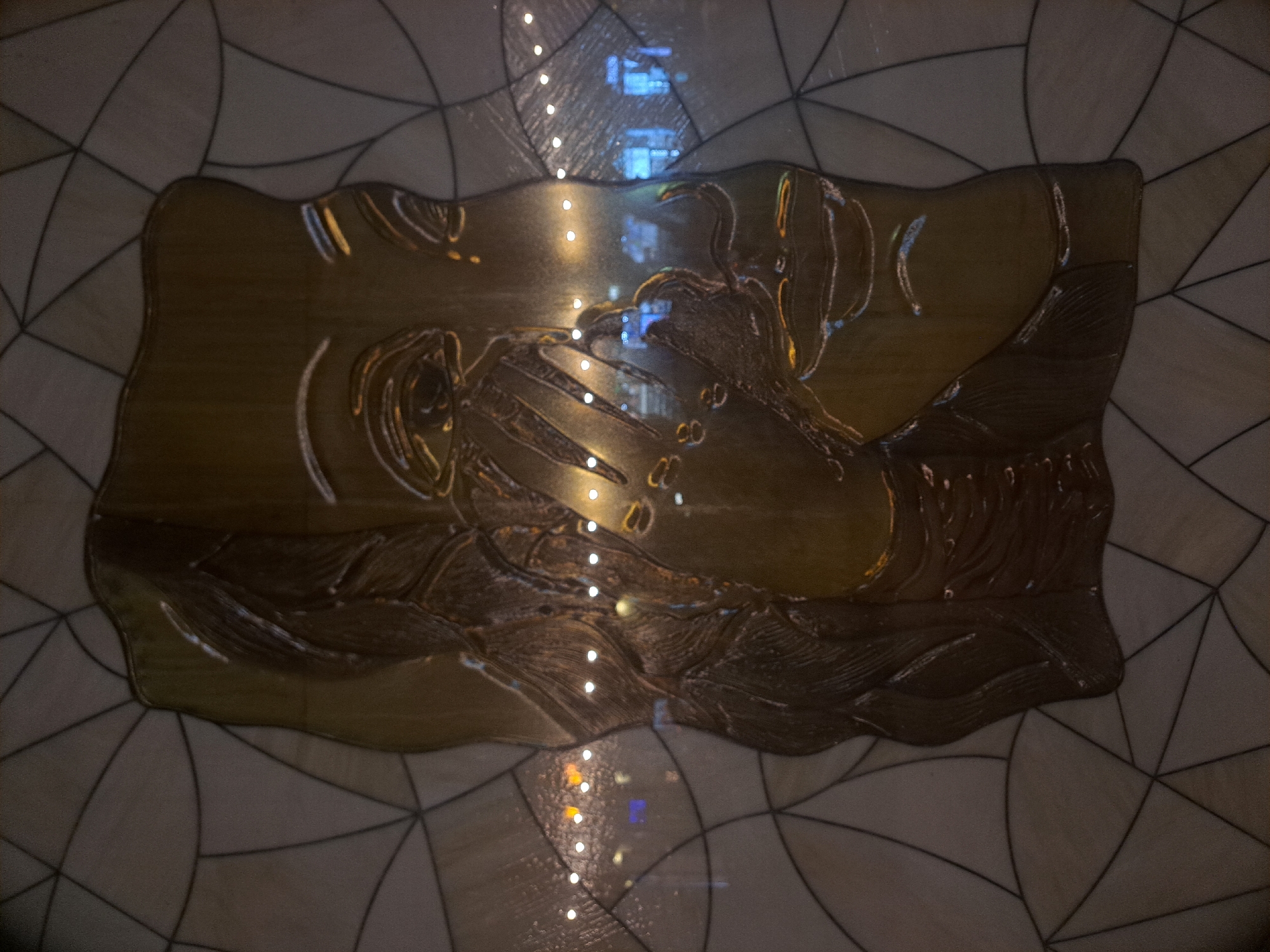 This is an image of a relief carving on a wall, likely indoors. The carving is rectangular with an irregular outline and depicts stylized figures. The carving is rendered in a golden-brown hue, possibly of metal or stone. A vertical string of light reflections runs through the middle of the image, suggesting it is capturing a reflective surface. The carving is set against a background of light-colored geometric patterns with dark lines, possibly a wall or floor tiling. The location is the Yerevan Cascade in Yerevan, Armenia.