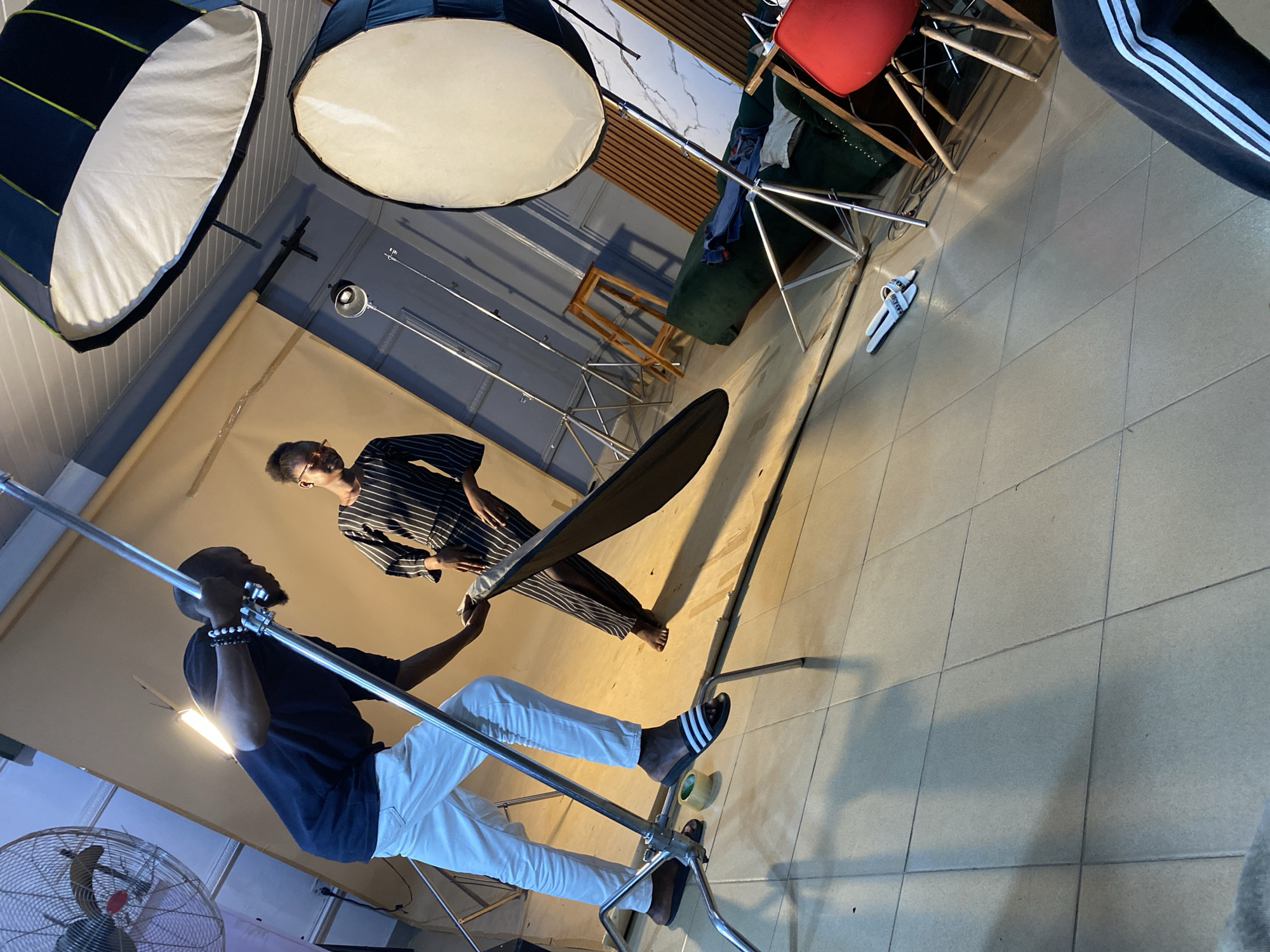 A person is holding a reflector and another person in striped clothing is standing in front of a backdrop. The environment includes studio lighting equipment, chairs, and other studio props. The location is Bukuru, Nigeria.