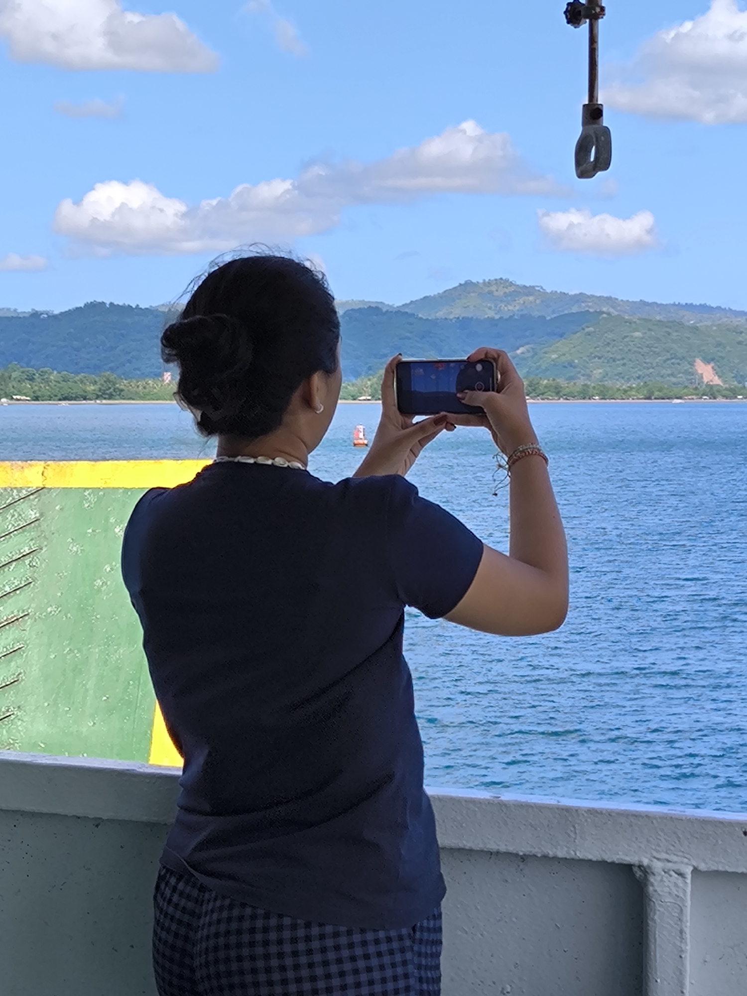 A daylight scene captured from the deck of a boat or ferry, showcasing a person taking a photograph of a picturesque seascape. The main subject is a woman, viewed from behind, facing away from the camera. She has dark hair tied in a bun and is wearing a dark blue t-shirt, a white shell necklace, and dark blue checkered pants. Her right wrist is adorned with a multi-colored bracelet.
She holds a black smartphone horizontally with both hands, aiming it at the horizon, indicating she is actively capturing the view. The phone screen shows a reflection of the landscape.
In the mid-ground, a wide expanse of calm, blue water stretches out, dotted with a small red and white buoy. The background features rolling green, forested mountains under a bright blue sky, scattered with fluffy white cumulus clouds. Parts of the vessel are visible in the foreground: a light gray railing, and sections of the boat's green and yellow painted metal structure on the left. Above, a metal bar with a hook-like attachment hangs down from the upper structure of the boat. The overall impression is one of travel and scenic appreciation in a coastal setting, likely in Lembar Selatan, Indonesia. No discernible text is visible in the image.