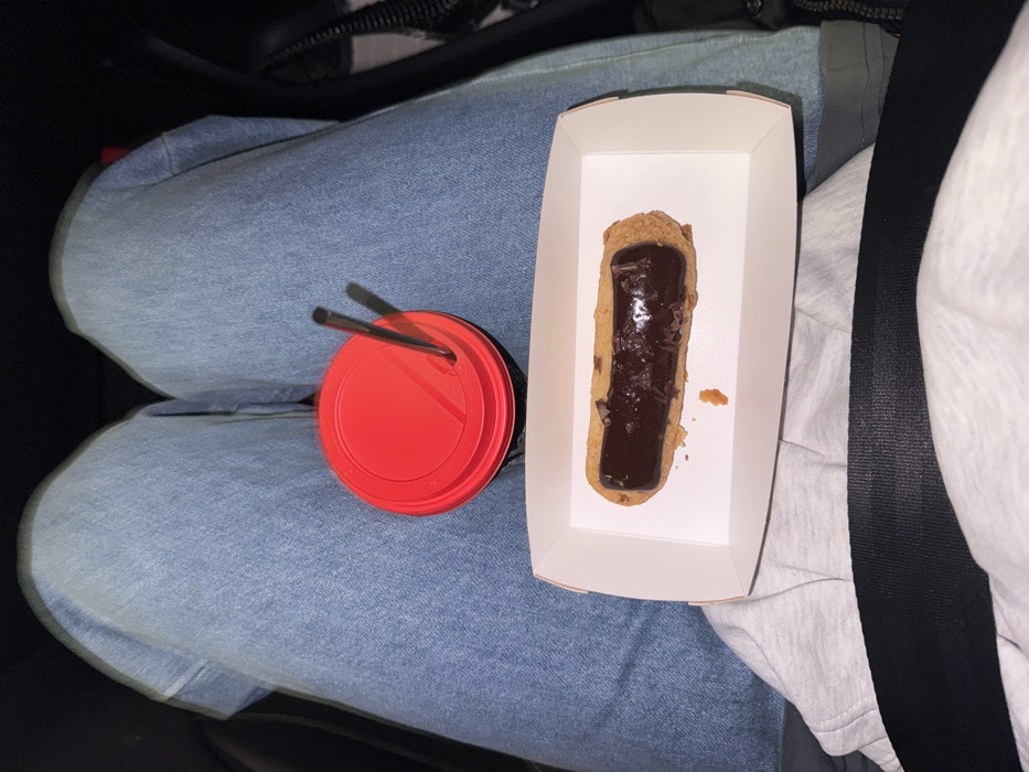 A red-lidded cup with two straws rests on blue jeans, next to a white cardboard container holding a chocolate éclair. A gray seatbelt strap lies adjacent to the container. The background is dark, hinting at a car interior.