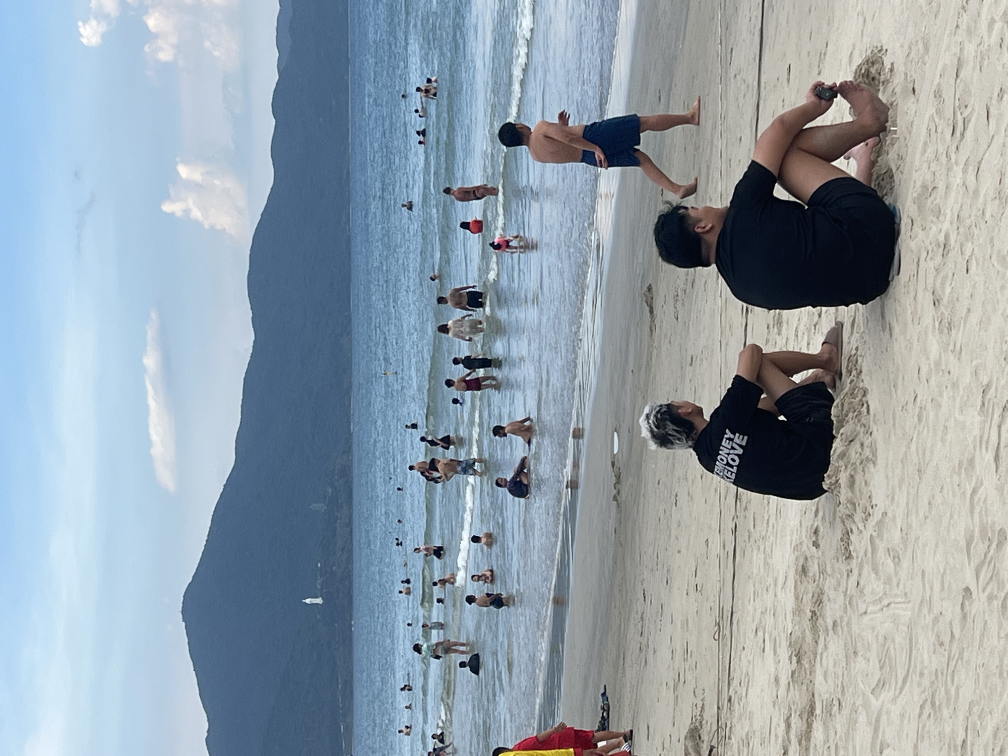 The image depicts a crowded beach scene in Đà Nẵng, Vietnam. The water is calm and shallow, with numerous people wading and swimming. The sand is light-colored and appears wet near the water's edge. In the background, a lush, mountainous landscape rises dramatically from the coastline, with some clouds dotting the clear blue sky. The sun appears to be high in the sky, suggesting it is midday.
The main subjects are the people enjoying the beach. Many are in the water, some standing, others swimming. On the sand, several individuals are visible, including two in the foreground. One person is sitting on the sand, wearing a black t-shirt with white text that reads "MONEY" and "LOVE" on the back, and black shorts. Another person is seated next to them, partially visible. Other people are scattered across the beach, some standing or walking.
The activity taking place is leisure and recreation at a beach. People are swimming, standing in the water, and relaxing on the sand. The atmosphere appears to be one of casual enjoyment and a pleasant day at the seaside. The overall impression is a typical summer day at a popular beach destination.