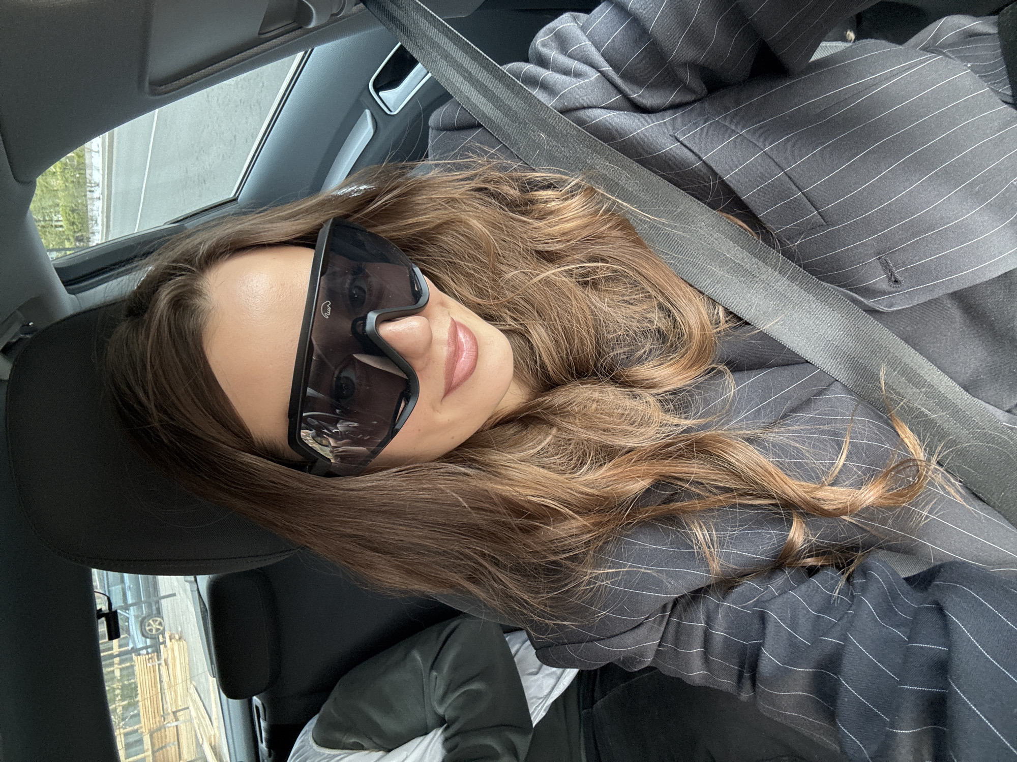 A woman with long brown hair, wearing oversized black sunglasses and a grey pinstripe suit, sits in the driver's seat of a car, secured by a seatbelt. The interior of the car is visible, including the headrest, sunroof, and side window. The background, seen through the window, shows buildings and greenery, possibly indicating a location in Krasnoyarsk, Russia.
