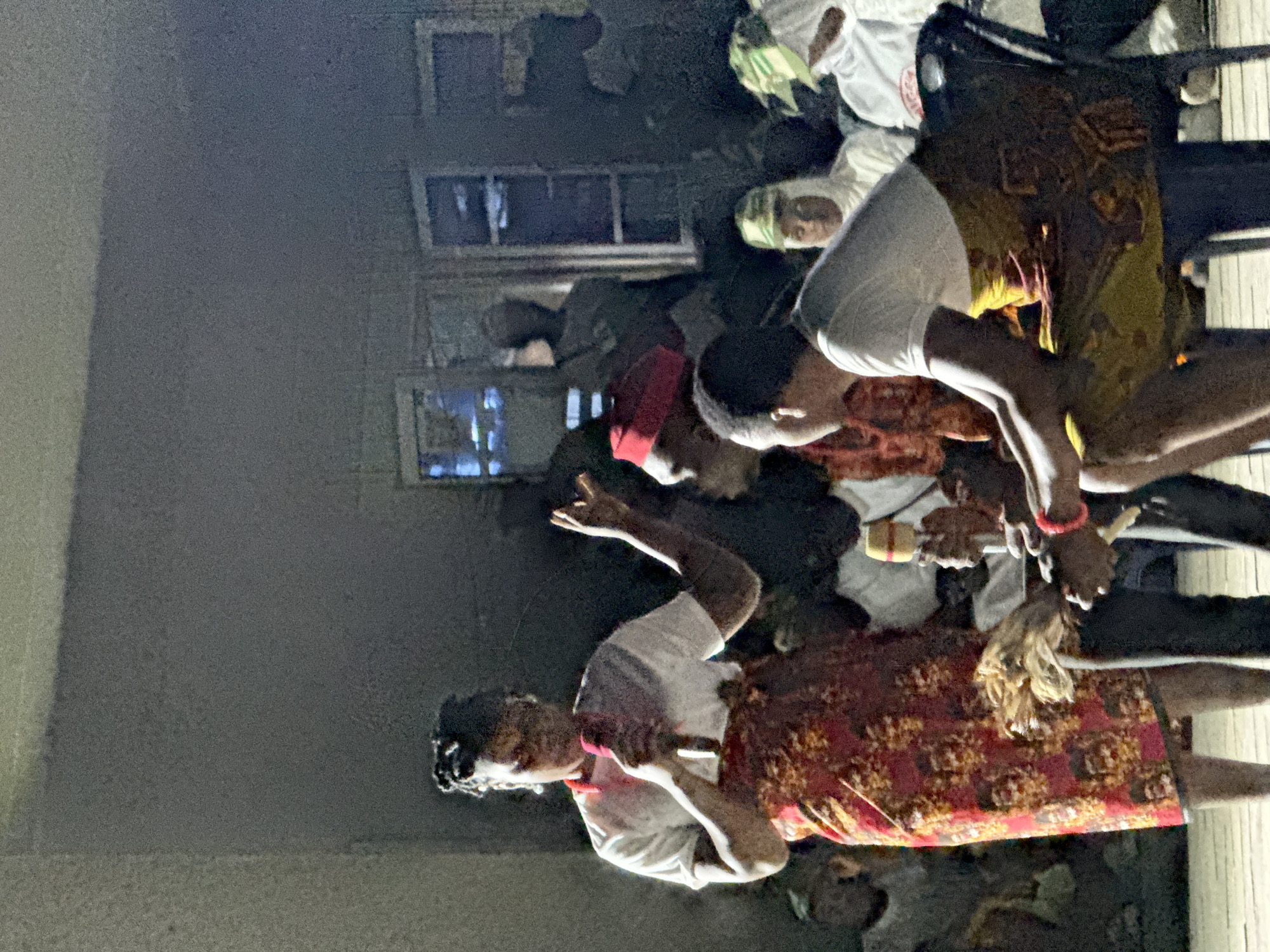 The image shows a group of people gathered outdoors at night. Some individuals are wearing traditional clothing, including patterned fabrics and head coverings. One person holds what appears to be a ceremonial object. The background includes a building with illuminated windows. The location is reported to be Maiduguri, Nigeria.