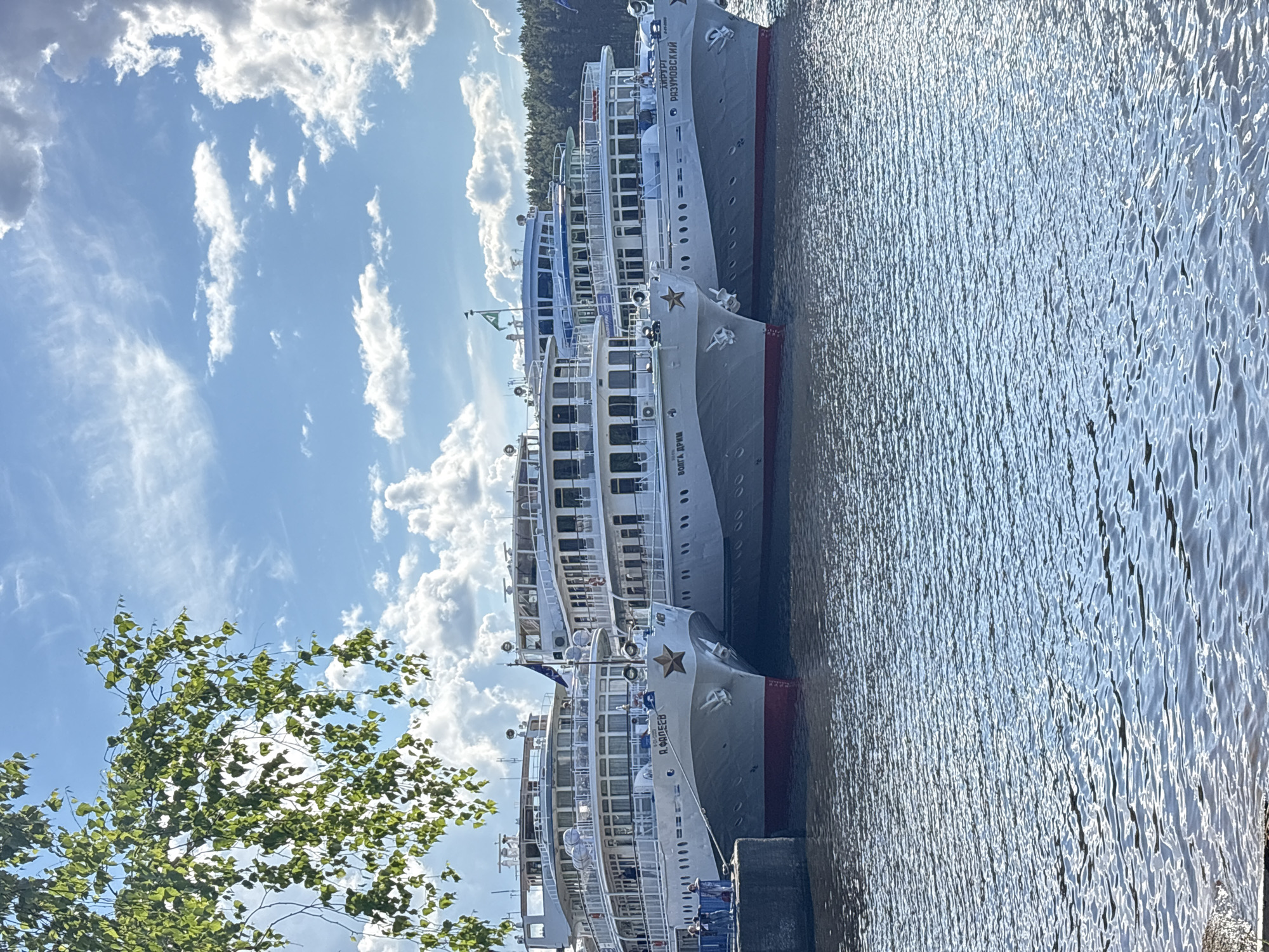 The image depicts a scene in the city of Chaikovsky, Russia, featuring several large passenger cruise ships docked along a waterfront. The ships are painted white and grey, with prominent red stars on their bows. Each ship has multiple decks and numerous windows. The water is calm and reflects the sunlight, creating a shimmering effect. The sky above is blue with fluffy white clouds. In the lower left corner of the image, a tree with green leaves is visible, suggesting a natural environment. A concrete pier or quay is present where the ships are docked.