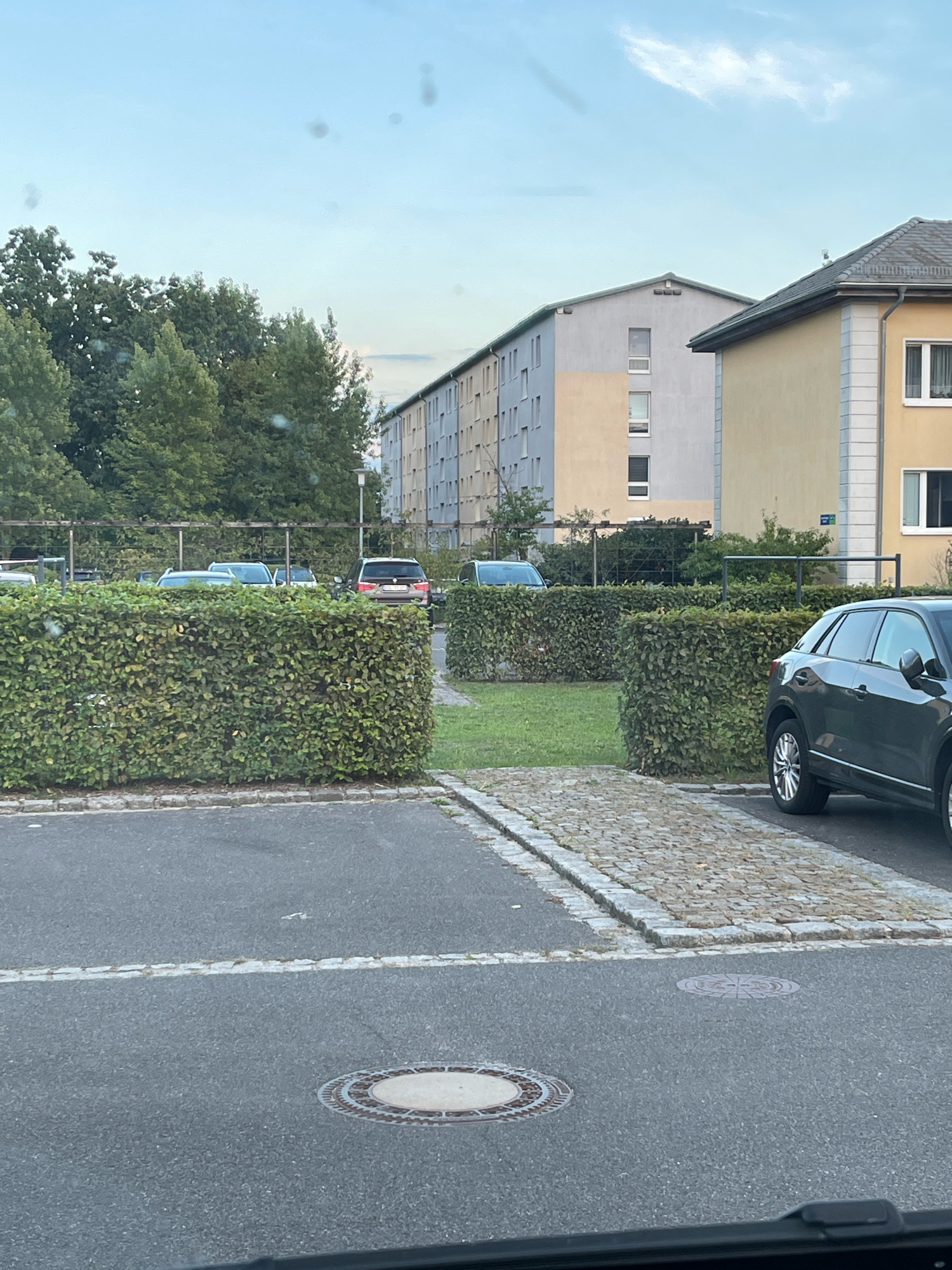 The image depicts an outdoor scene in the city of Lübbenau/Spreewald, Germany. In the foreground, a dark-colored SUV is parked on a paved area with cobblestone accents. Beyond the SUV, a well-maintained hedge forms a barrier, separating the parking area from a grassy space. In the middle ground, several other vehicles, including SUVs, are visible parked behind the hedge. A long, multi-story building with a light-colored facade and numerous windows runs along the background. Adjacent to this building is another structure with a yellow facade and a grey tiled roof. The sky above is partly cloudy with a pale blue hue. Trees with dense green foliage are present on the left side of the image, bordering the buildings. The overall impression is a residential or apartment complex with designated parking areas.