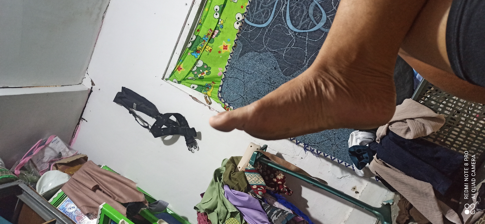 In Sedati Agung, Indonesia, a close-up perspective shot shows a person's foot and lower leg against a cluttered background. The foot is prominently positioned in the foreground, with its sole facing the camera and toes slightly curled. The skin texture is visible, showing wrinkles and creases. The background is filled with various objects, including piles of folded clothes in earthy tones like beige and brown, as well as darker fabrics like navy blue and purple. A green object, possibly a tool or a piece of equipment with a long handle, is leaning against the wall. Further back, a black strap with buckles is attached to the white wall. To the right, a green fabric with a cartoon frog print covers a surface, possibly a bed or a mattress. A white water bottle cap is also visible among the clutter. The lighting suggests an indoor environment, possibly a room in a home, with light reflecting off the objects.