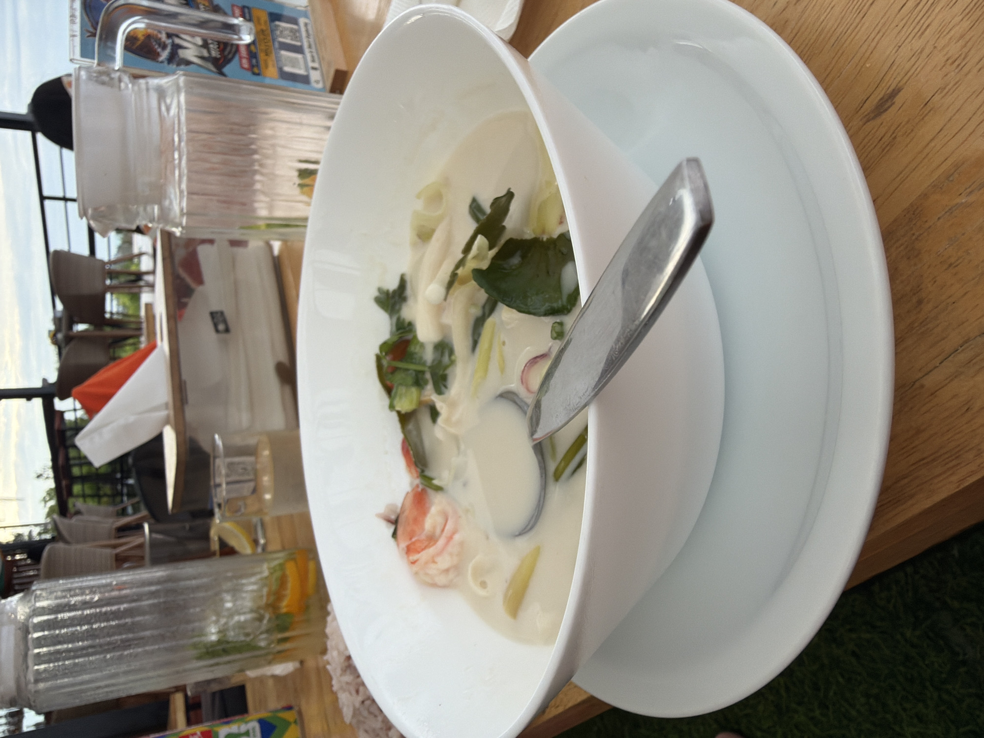 In Ko Samui District, Thailand, a close-up shot captures a bowl of Tom Yum soup on a wooden table. The soup is creamy white, with visible pieces of shrimp, mushrooms, and herbs. A metal spoon rests in the bowl. To the left of the soup, a tall glass pitcher filled with water, sliced oranges, and mint is present. To the right, another glass pitcher contains clear water. In the background, a blurred scene shows an outdoor seating area with chairs and tables, suggesting a restaurant setting. The lighting indicates it is daytime.