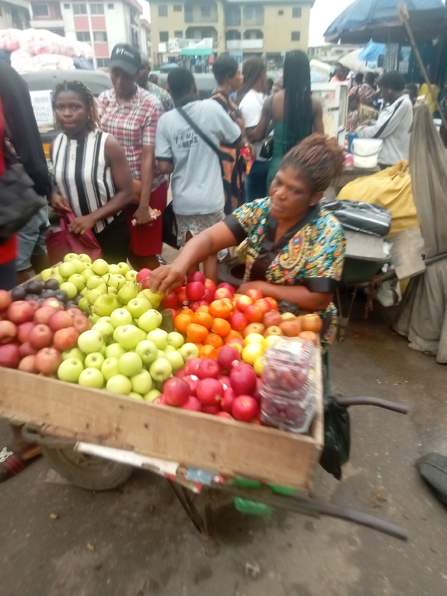 In a crowded street market in Onitsha, Nigeria, a woman wearing a colorful patterned top stands behind a wooden cart filled with various fruits: green and red apples, oranges, and grapes. She is selecting a red apple. Around her, several people are walking or standing nearby. The buildings in the background suggest an urban setting. The ground is paved, appearing somewhat worn. Umbrellas and canopies from other vendors are visible in the distance.