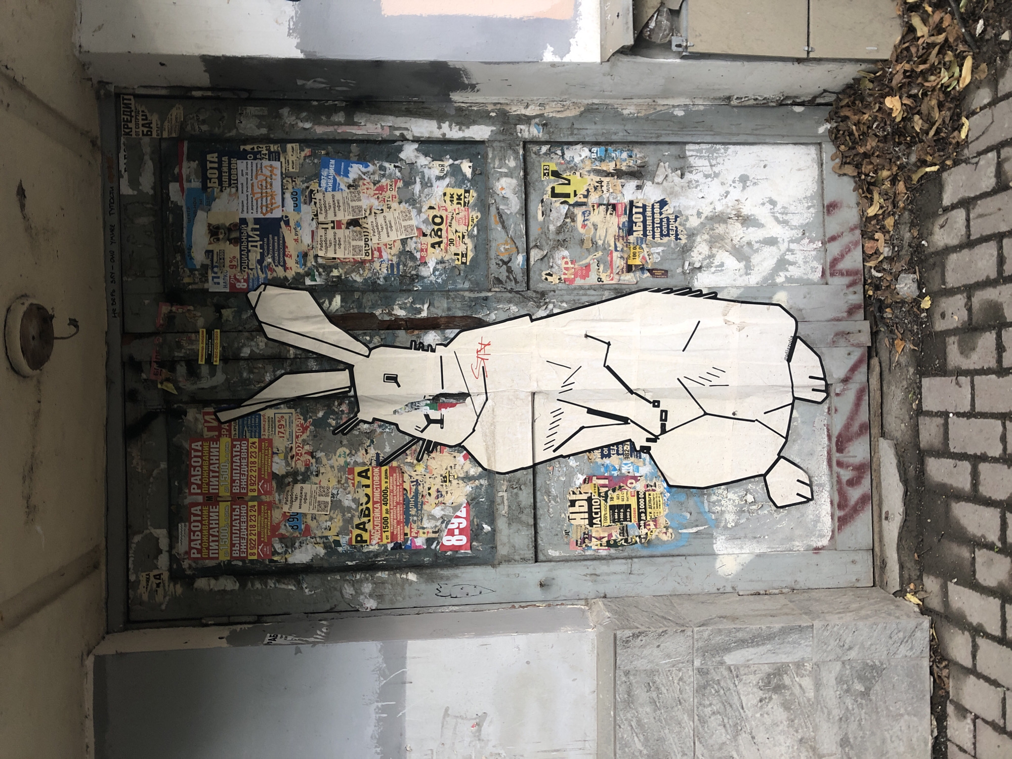 The image shows a weathered metal door covered in numerous posters and advertisements, with a large, stylized white rabbit graffiti drawing superimposed on it. The door is situated within a building's entrance, flanked by grey stone-tiled walls. The street in front of the door is paved with brickwork, and a pile of fallen autumn leaves is visible at the base of the door. The graffiti art is a minimalist depiction of a rabbit, outlined in black, with simple geometric shapes forming its body. Some of the posters are faded and torn, suggesting a public space with high foot traffic. Based on the poster content, which includes job advertisements and financial offers in Russian, the location is likely Yekaterinburg, Russia. The overall impression is one of urban decay and street art.