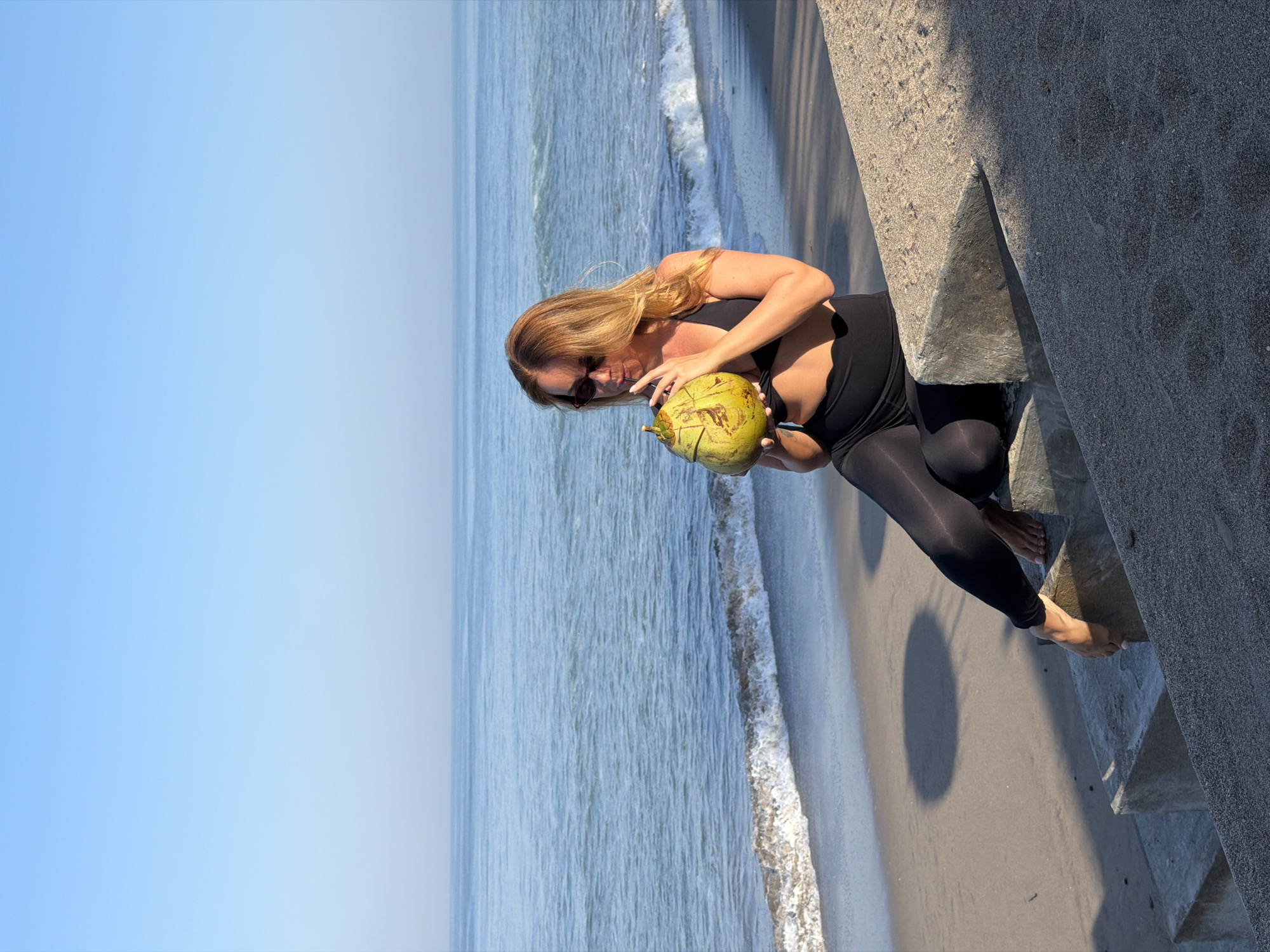 A woman is sitting on concrete steps by the beach in North Kuta, Indonesia. She is wearing a black sports bra and black leggings, and sunglasses. She is holding a young green coconut and drinking from it with a straw. The ocean is behind her with waves breaking on the shore. The sky is clear and blue.