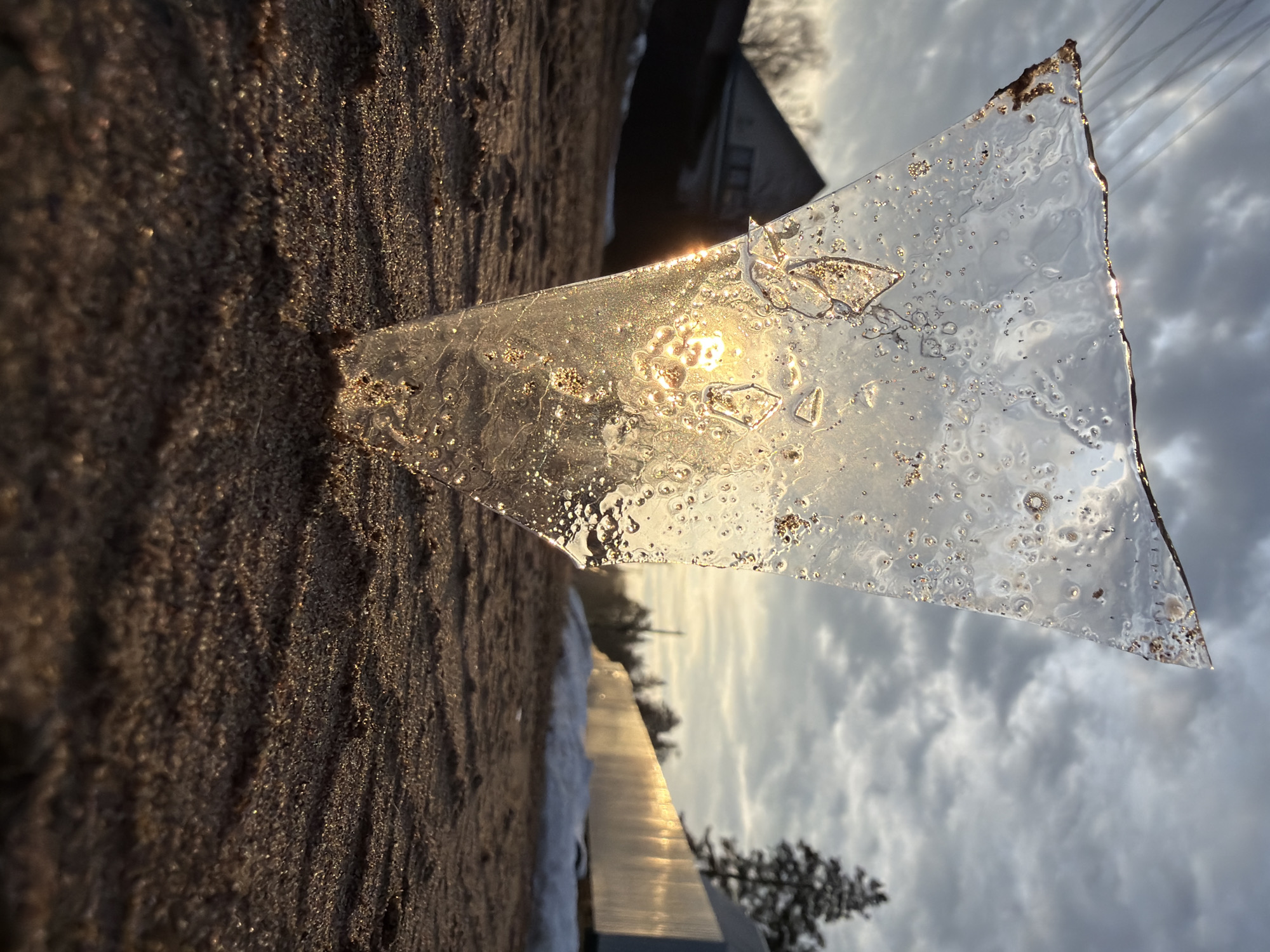 A low-angle, outdoor shot depicts a triangular piece of ice partially embedded in a sandy surface, with the sun shining through it. The ice appears translucent, with small air bubbles and particles trapped within. The sandy surface is coarse in texture, with varying shades of brown.
In the background, partially obscured by the ice, a cloudy sky is visible, along with distant trees and a building. A metallic or reflective object is also partially visible, suggesting it might be a part of a building or structure. The location is identified as Chaikovsky, Russia, though specific details within the image to confirm this are not apparent.
