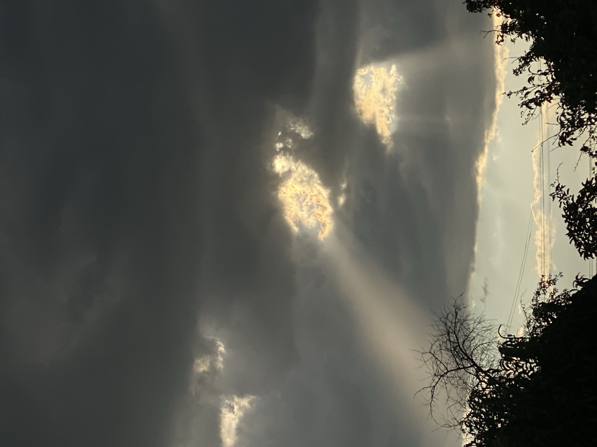 The image depicts a dramatic sky with dark, ominous clouds pierced by shafts of sunlight. In the foreground, silhouetted against the lighter sky, are the tops of trees and branches. The scene is likely captured during twilight or a storm. The location is identified as Cairaclia, Moldova. There are no people or overt actions visible in the image. The environment is characterized by the interplay of light and shadow in the sky and the dark, natural forms of the trees.