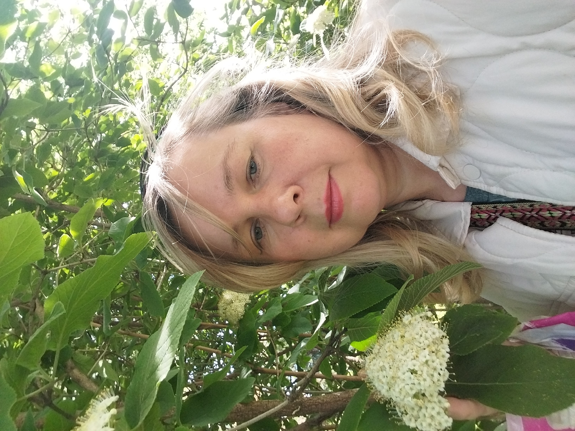 A blonde woman wearing a white jacket is positioned among green foliage and clusters of small white flowers. The image is oriented at an angle, with the woman's face tilted towards the camera. The setting appears to be outdoors, surrounded by a dense collection of leaves and branches. There's no clear indication of the location being the city of Orenburg, Russia within the picture itself.
