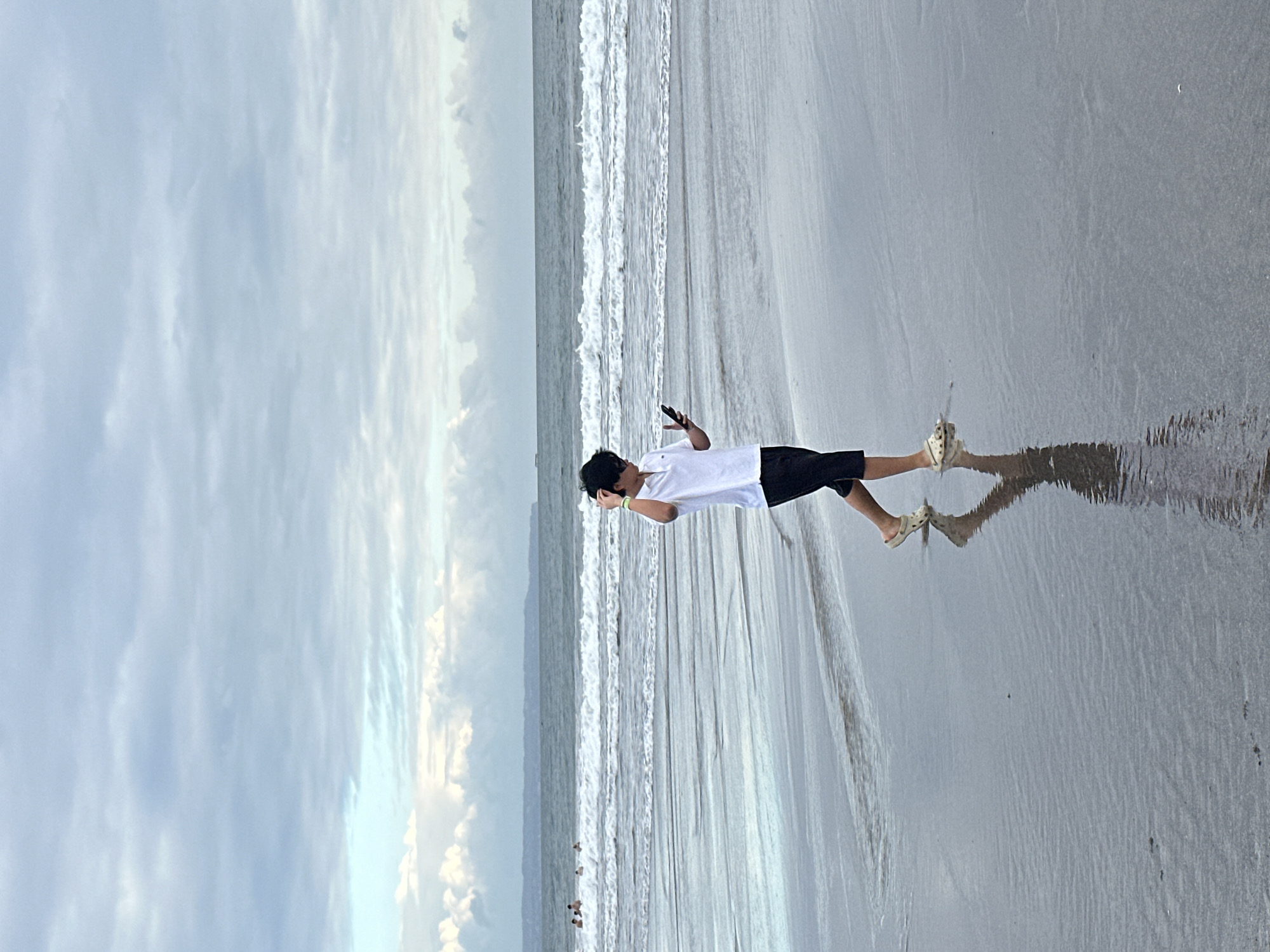 A lone individual is walking on a beach in North Kuta, Indonesia. The individual is wearing a white polo shirt and black shorts, with light-colored slip-on shoes. They are looking at their phone and have their hand near their head. The beach has wet sand that reflects the sky. In the background, there are gentle waves rolling onto the shore and a few other people in the water. The sky is overcast with a mix of gray and white clouds. The overall scene suggests a calm, somewhat hazy day at the beach.