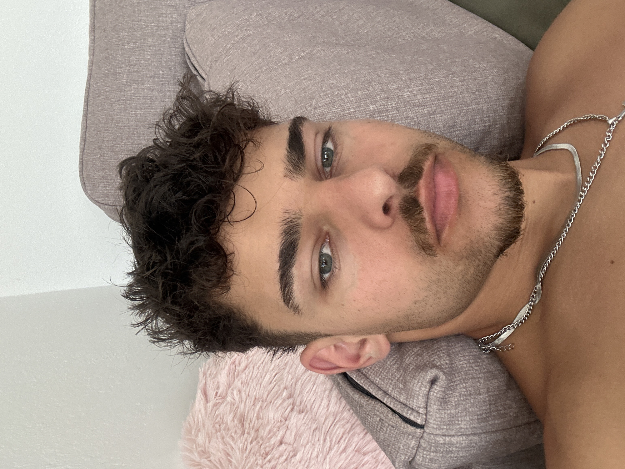 A medium shot of a man lying on a grey cushion, possibly a sofa. He has short dark curly hair, light green eyes, and facial hair including a mustache and goatee. He is wearing a silver chain necklace. The background includes a white wall and a pink fuzzy pillow. The location is not discernible from the image; no information indicates this image was taken in Troyan, Bulgaria.