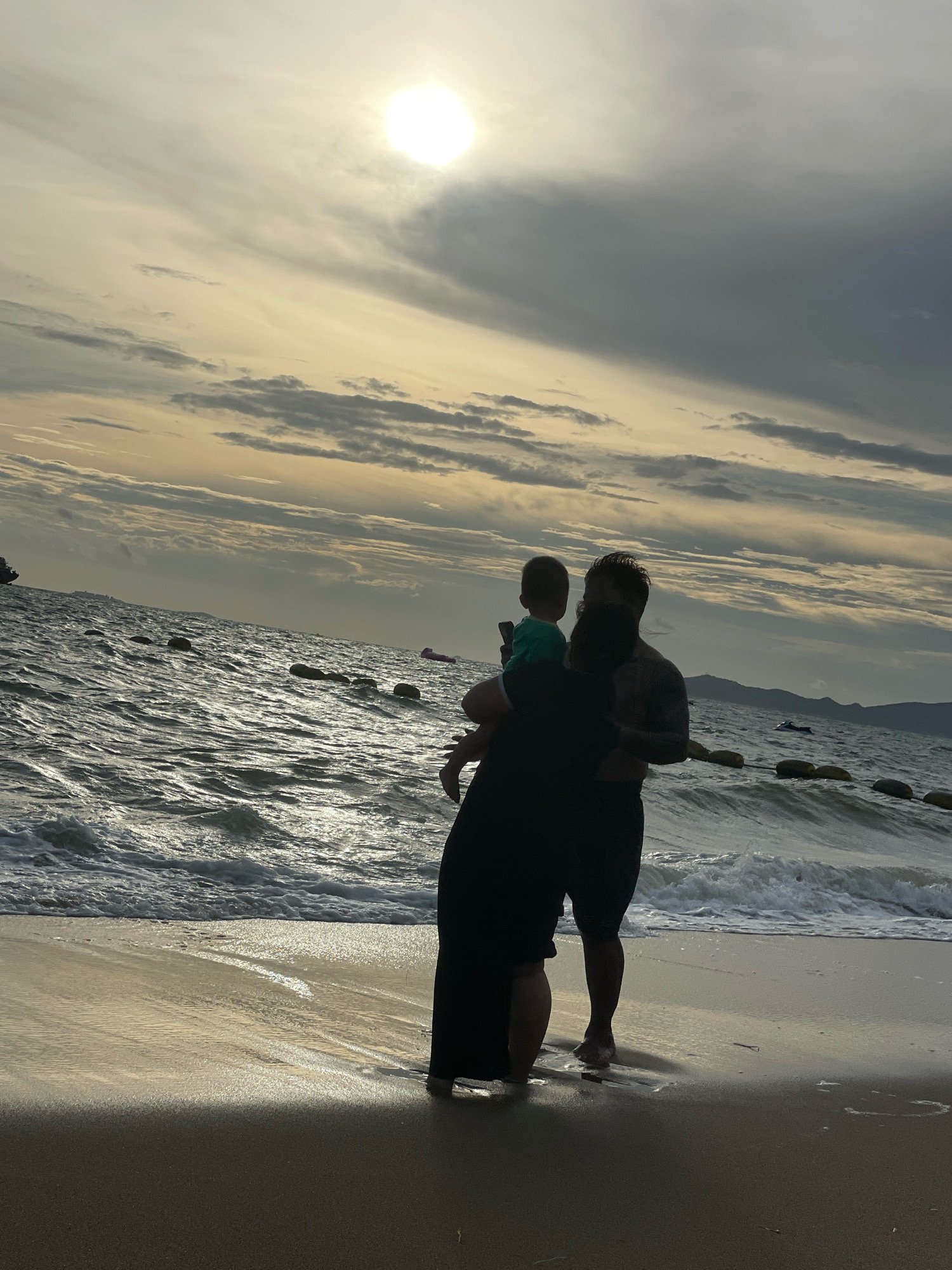 A family of three, consisting of two adults and a child, stand on a sandy beach facing the ocean at sunset in Pattaya City, Thailand. The adults are silhouetted against the sky, with the taller adult carrying the child. The child is facing away from the camera, looking out at the sea. The ocean is choppy, with waves breaking on the shore. The sky is filled with dramatic clouds, with the sun partially obscured by them. In the distance, there are mountains and a jet ski. Yellow buoys are visible in the water, marking a swimming area. The sand is wet from the receding tide, reflecting the light of the setting sun. The overall scene is peaceful and evokes a sense of family bonding and the beauty of nature.