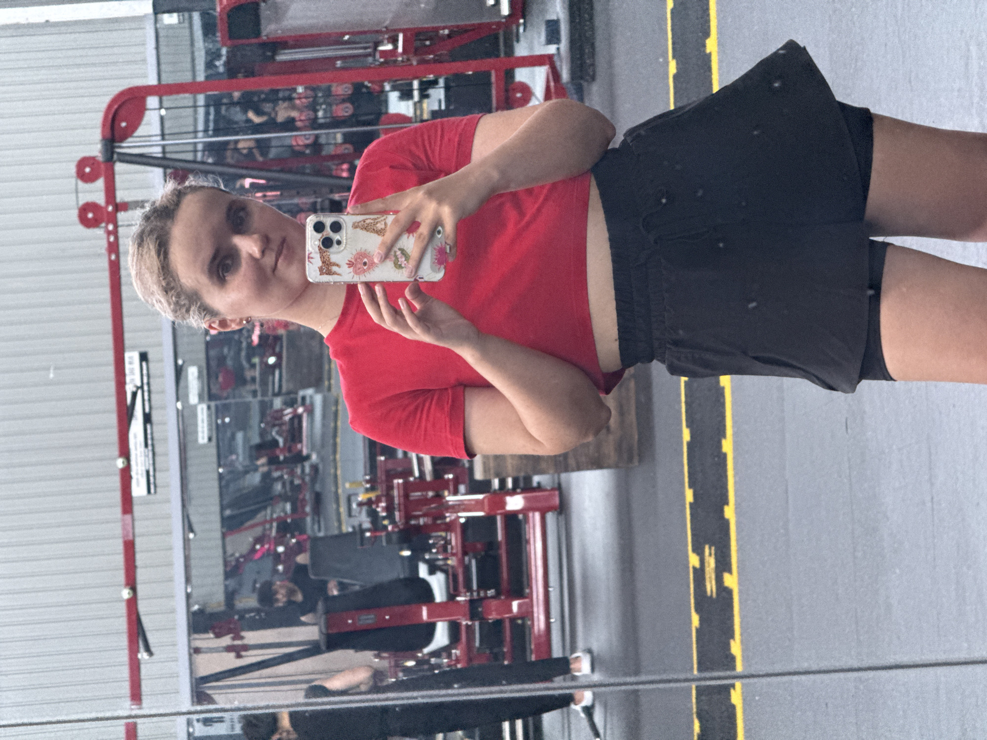 A young woman is taking a selfie in a gym. She is wearing a red crop top and black shorts. The gym equipment is visible in the background, including a weight machine. The location is identified as Da Lat, Vietnam based on the gym's specific architectural style. The floor is gray with a black and yellow caution stripe. She is holding a smartphone in her right hand and facing the mirror.