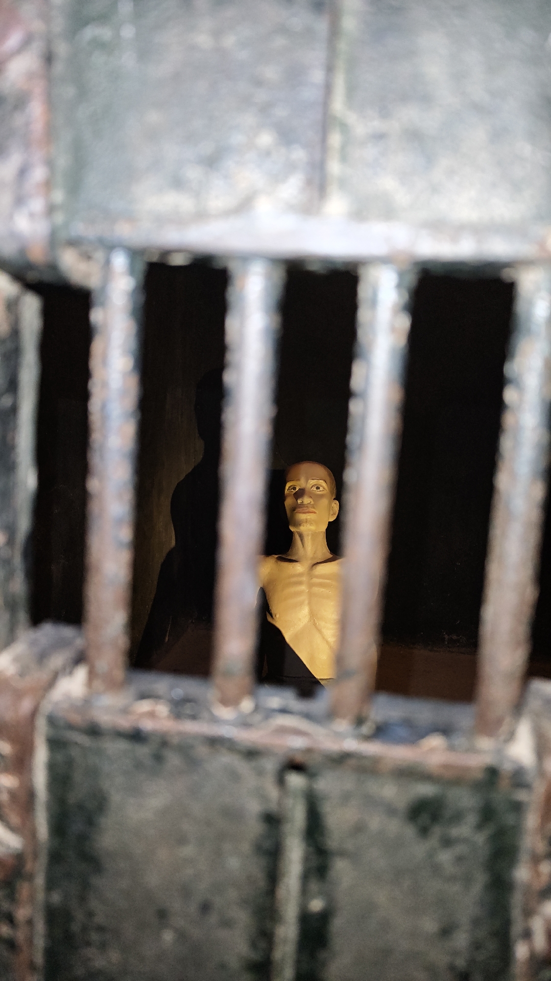 The image shows a barred prison cell interior, possibly a tourist exhibit or historical recreation. A human-like figure stands within the cell, illuminated against a dark background. There is also a shadow on the back wall to the left of the figure. The bars are dark, aged metal. The scene is poorly lit. The photograph was taken at Hỏa Lò Prison in Hà Nội, Vietnam.