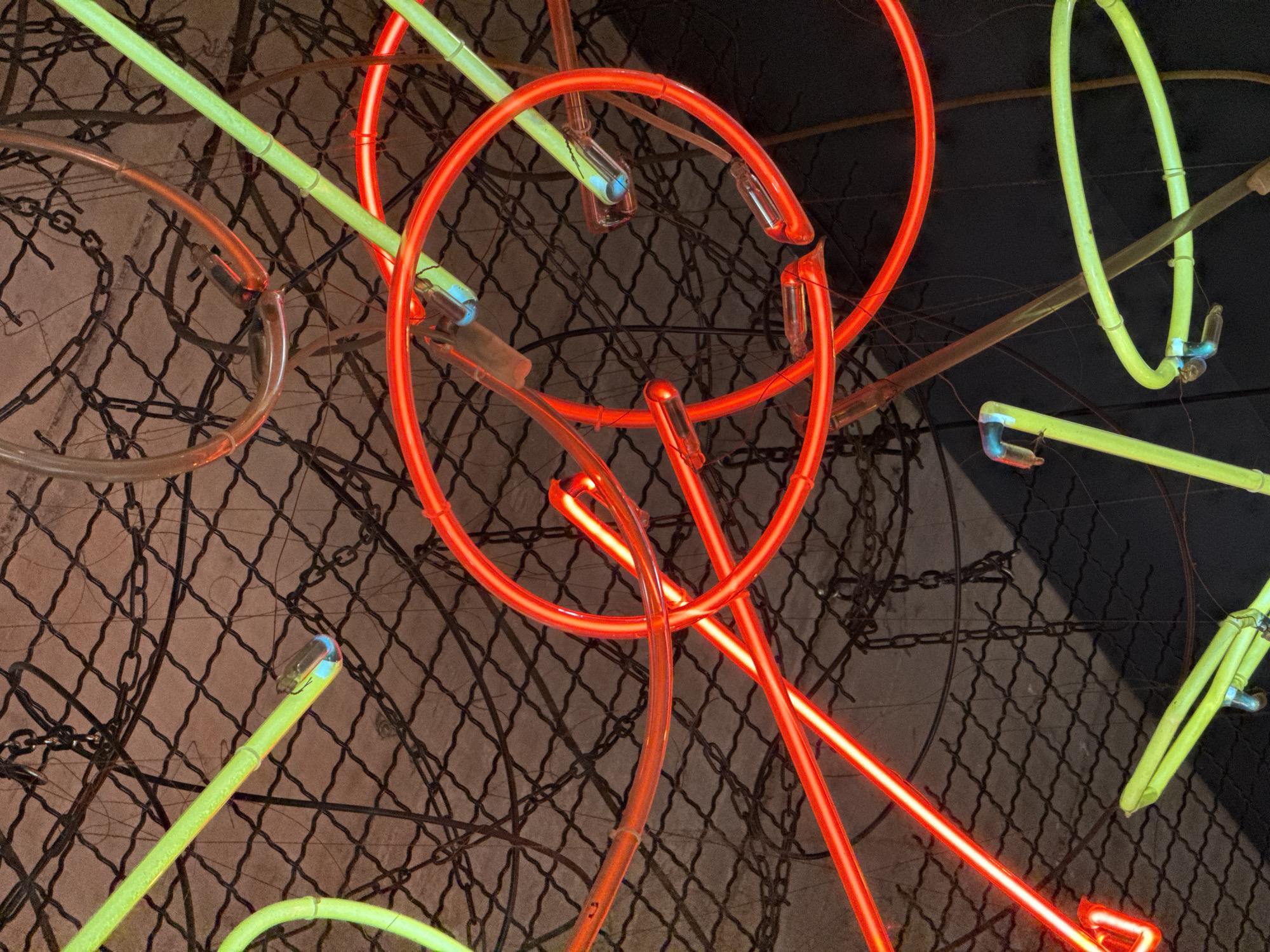 The image displays an abstract artistic arrangement of illuminated neon lights against a dark, textured background. The primary subjects are vibrant neon tubes, prominently featuring bright red and a distinct light green or chartreuse color. These tubes are shaped into various curves, circles, and straight lines, overlapping and intersecting to create a dynamic composition. Visible glass connectors and electrodes at the ends of some tubes emit a faint blue light.
The background consists of a dark, grid-like pattern resembling a chain-link fence or wire mesh. Several dark, metallic chains are also discernible, draped or interwoven within this background, suggesting an industrial or urban setting. The overall scene implies a static art installation or display.
The strong glow of the neon lights indicates either a nighttime environment or a very dimly lit indoor space, allowing their colors to dominate. No people or overt activity are present. The image conveys a modern, energetic, and somewhat raw aesthetic due to the combination of glowing artificial light and the gritty backdrop. There is no visible text. The location context provided for this scene is Podgorica, Montenegro.