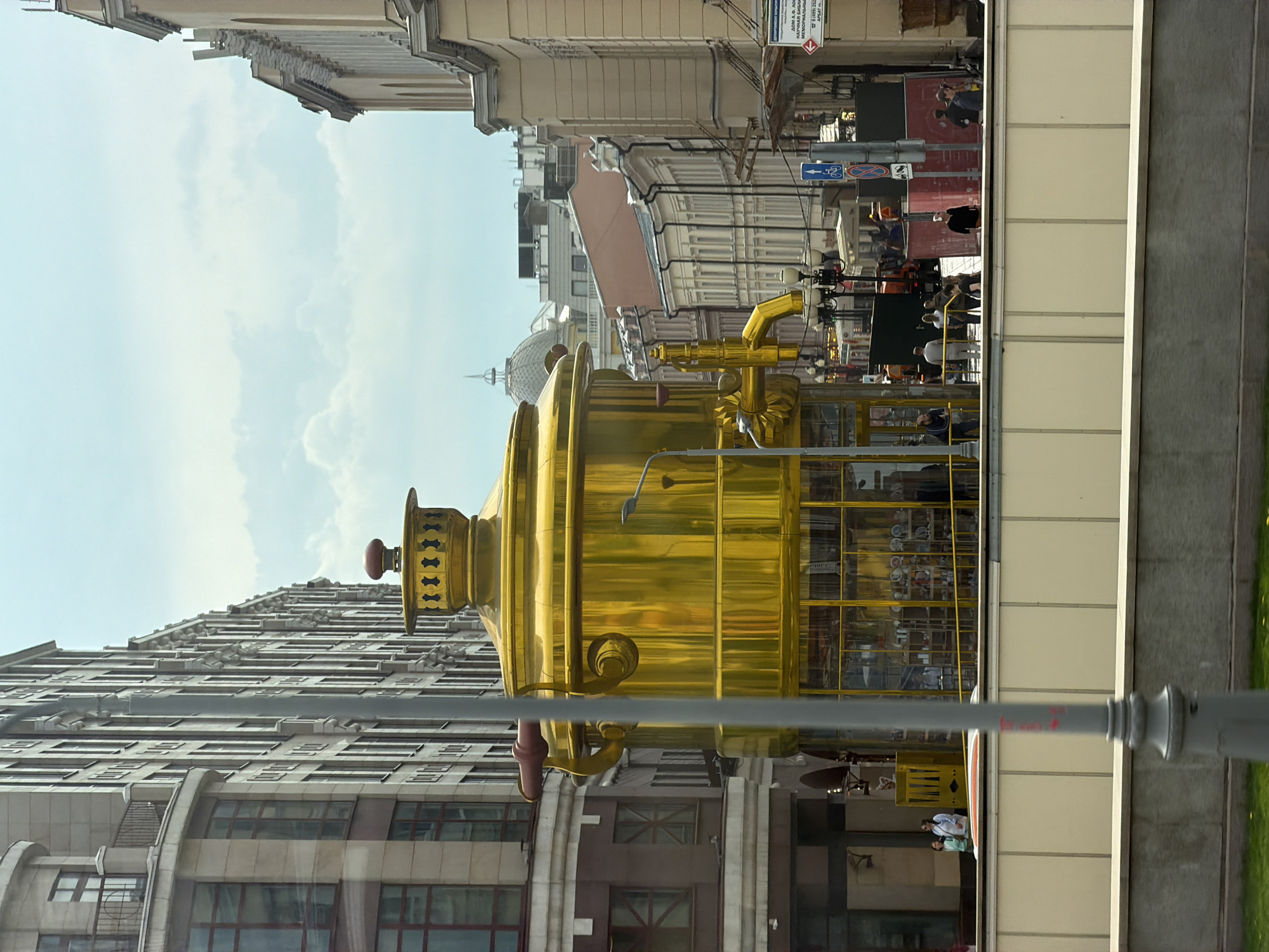 This photograph presents a scene in Moscow, Russia, featuring a large, golden samovar structure on a street with mixed architectural styles. The imposing samovar, gleaming with a high-gloss finish, is positioned in a way that interacts with the surrounding cityscape, creating a somewhat surreal juxtaposition against the more traditional buildings.
The environment includes modern architectural buildings with multiple floors and repetitive window patterns, as well as older buildings in the background, suggesting a blend of old and new within the urban fabric. The presence of street signs, pedestrian traffic, and a public transportation vehicle (possibly a bus or tram) indicate an active city atmosphere.
The people are seen interacting within the scene, both inside and outside of what appears to be a public transportation structure. There is also a streetlamp visible in the foreground. The interaction between these various elements forms a dynamic urban scene.