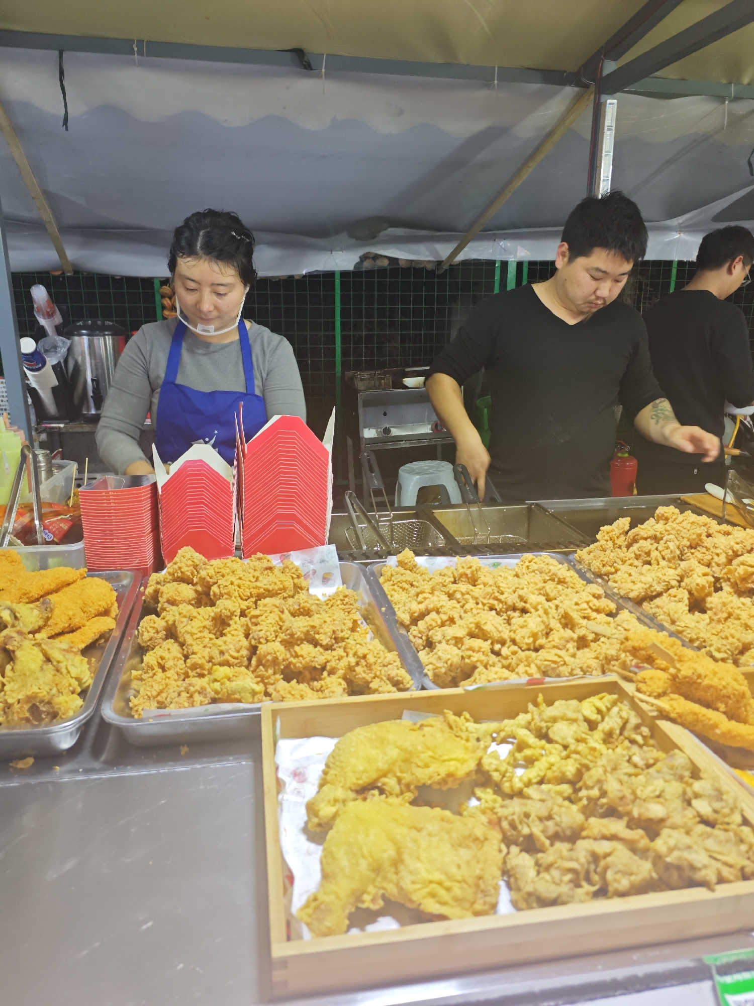 The image shows a food stall with three people working, likely in Shapingba District, China. In the foreground, there are several trays filled with fried food, including what appears to be fried chicken and other battered items. Red takeout boxes are stacked on the counter. A woman wearing a blue apron and a face shield is working behind the counter, attending to the food. A man in a black shirt is using tongs to manage food in a deep fryer. A third person, also in a black shirt, is partially visible in the background on the right side of the frame. The stall is lit by overhead lights. The background reveals a green fence.