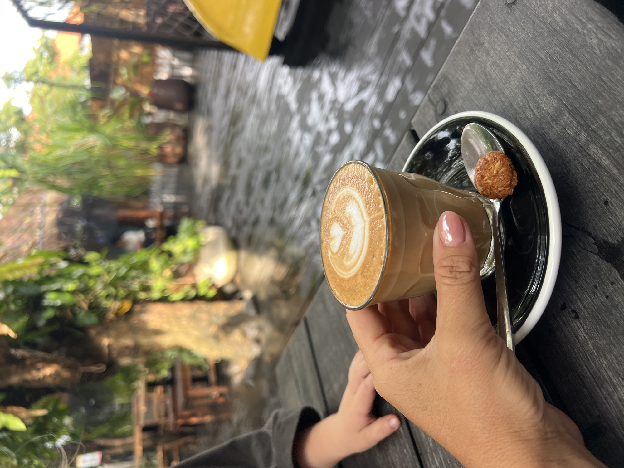 A hand holds a glass of coffee with latte art on a saucer next to a spoon and a small, round pastry. The coffee is in a clear glass cup and appears to be a macchiato or flat white. The pastry is small and textured, possibly a cookie or confection. The scene is set outdoors on a dark wooden table. In the background, there is a child's hand, lush greenery, and wooden furniture, suggesting an outdoor cafe or restaurant setting in North Kuta, Indonesia. The ground appears wet, indicating recent rain. The overall impression is one of a relaxed, casual moment.