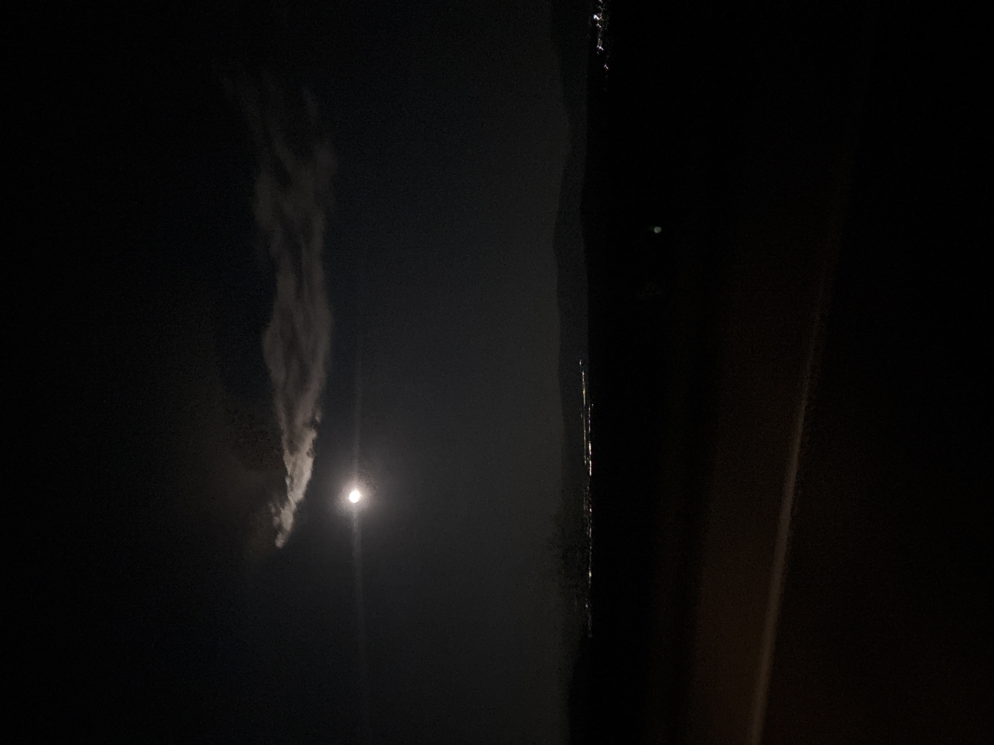 The image appears to be taken at night. A bright light source, likely the moon, is partially obscured by a cloud. To the right of the light source is a dark, vertical structure that could be a building or a large object. The overall environment is dark and it is difficult to make out details. There are no visible people, actions, or interactions in the image. Given the prompt mentioning the location, the picture could be taken in Lesichovo, Bulgaria.
