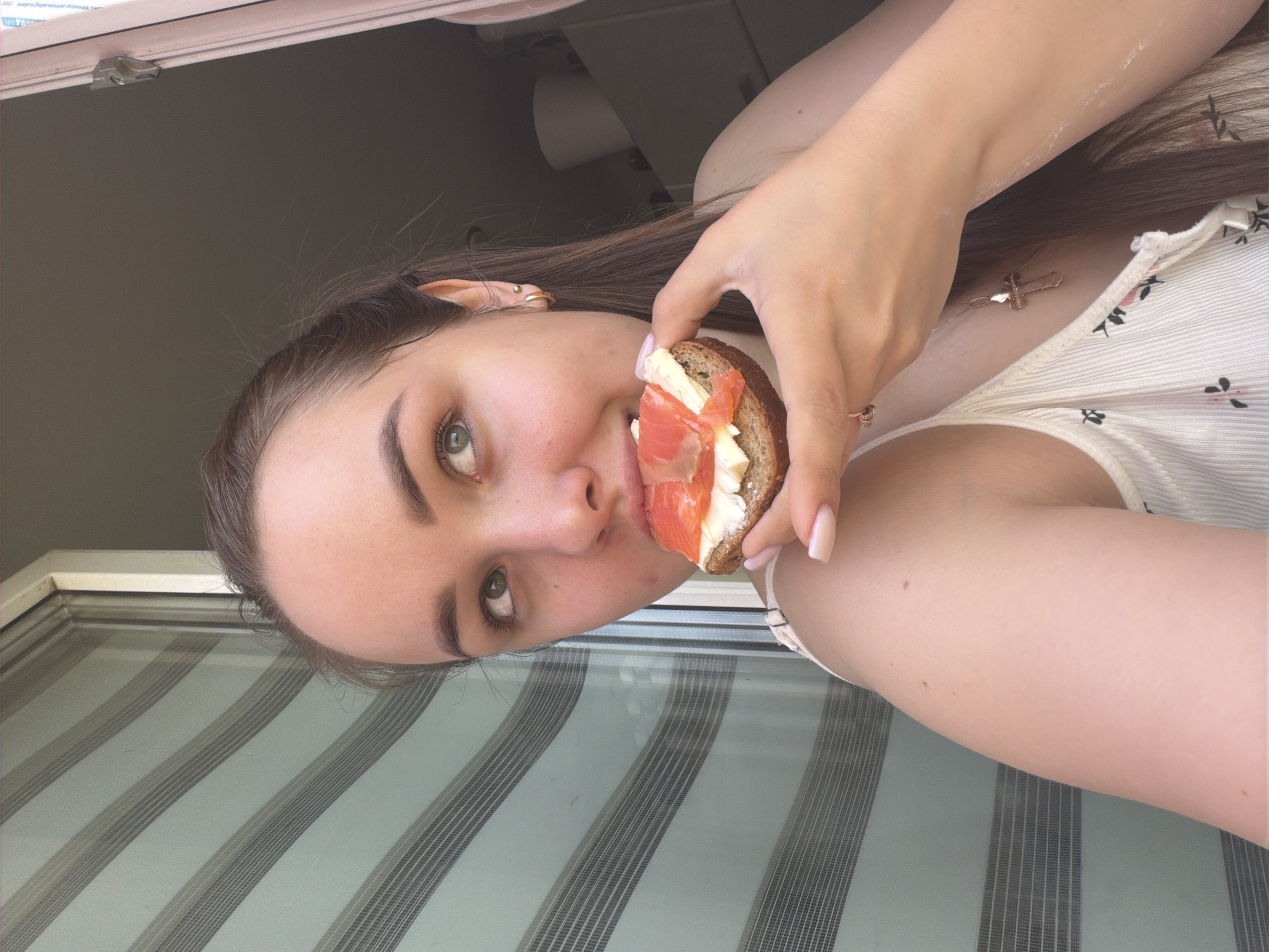 A young woman is holding a sandwich up to her mouth, likely about to take a bite. The sandwich appears to be topped with salmon and cheese. The woman is wearing a light-colored top. The background features a window with blinds. The location is Chaikovsky, Russia.