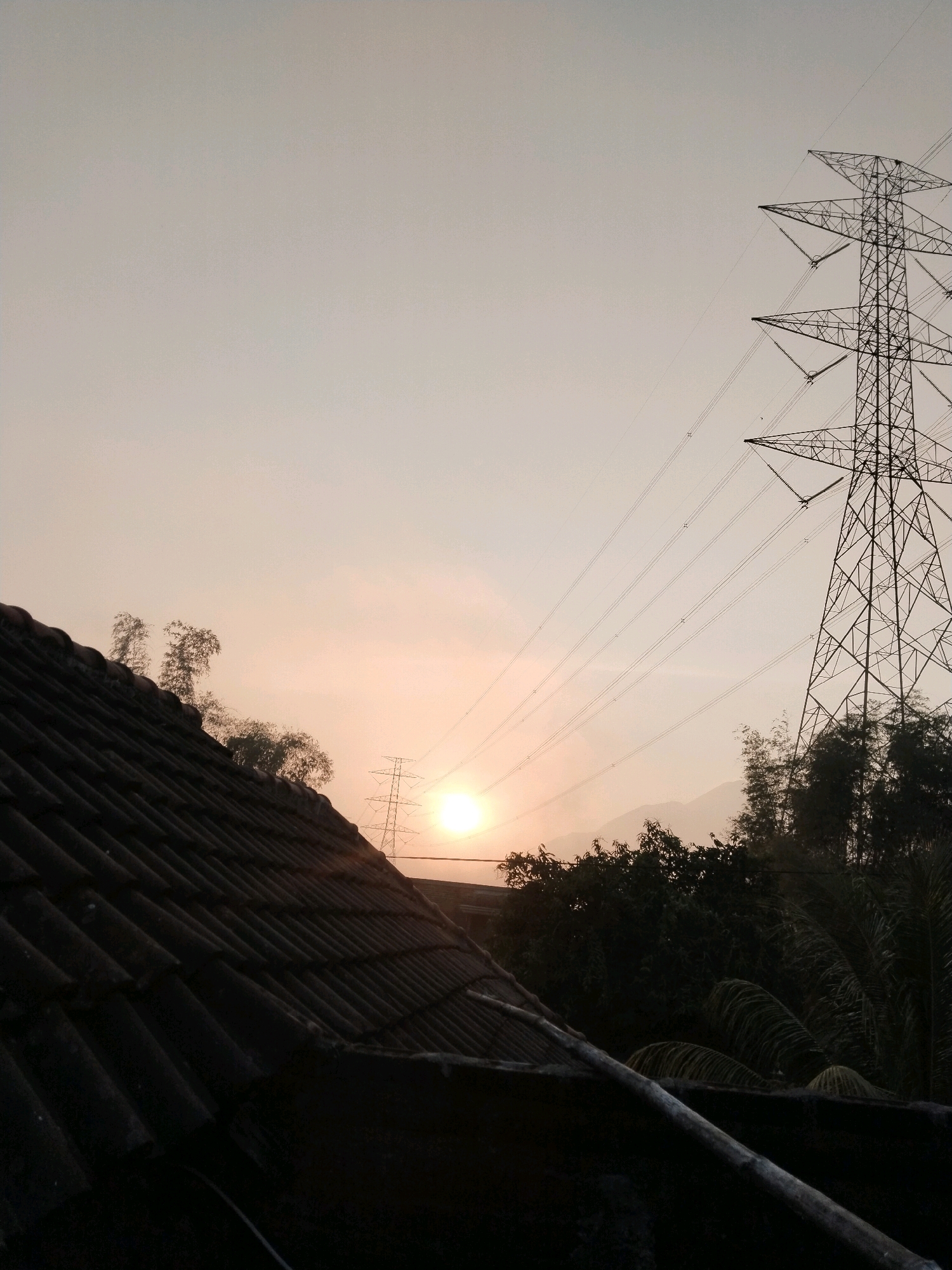 The image depicts a sunset scene viewed from a rooftop in Kuripansari, Indonesia. In the foreground, the dark, tiled roof of a building occupies the lower left portion of the frame. Beyond the roof, the sky is a soft, hazy orange and yellow, illuminated by the setting sun, which is partially obscured by a distant transmission tower. Several power lines stretch across the sky from a large, lattice-style transmission tower located to the right of the frame. Silhouetted trees and foliage form a dark band across the middle ground, with distant mountains faintly visible against the horizon. The overall impression is one of a serene, natural landscape juxtaposed with industrial infrastructure.