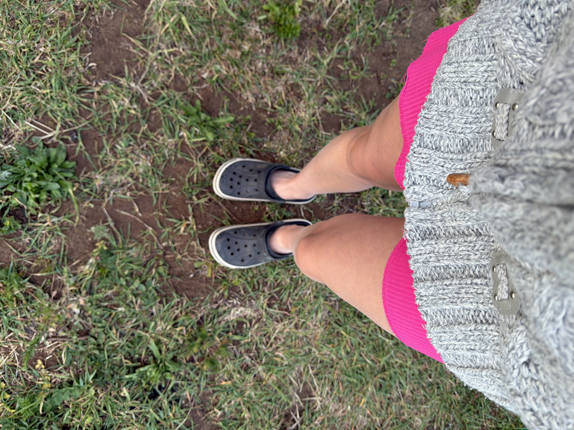 A person wearing black Crocs, a pink skirt, and a grey sweater stands on a patch of grass and dirt. The location is Chaikovsky, Russia.