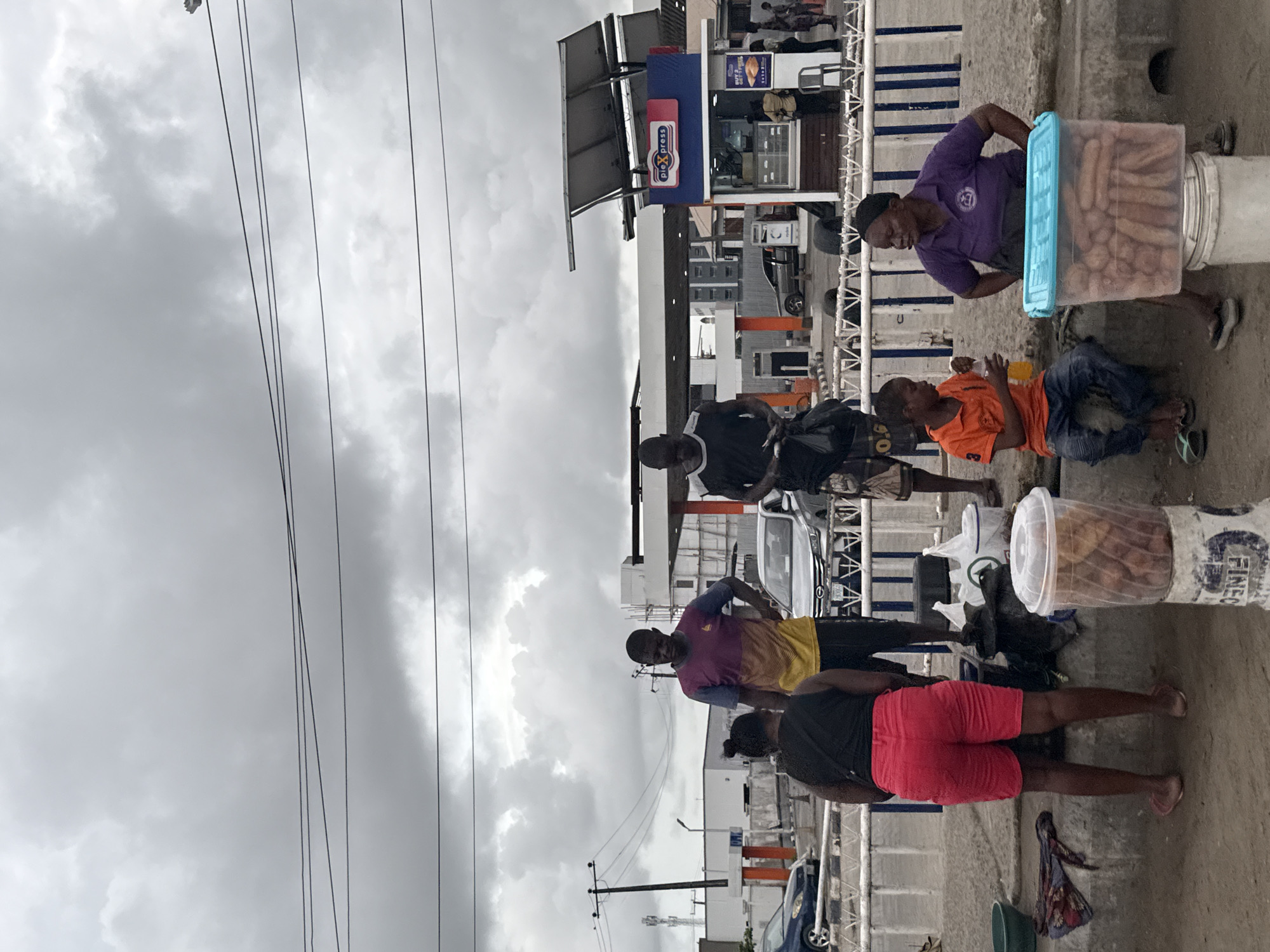 The image, taken during daytime under an overcast sky, captures a street scene likely in Lekki, Nigeria, given the provided context. It features several individuals engaged in what appears to be street vending activities near a commercial establishment.
In the foreground, a man in a purple short-sleeved shirt stands beside a large, transparent plastic container filled with fried snacks, possibly puff-puff or similar, which rests on a white bucket. Next to him, a child in an orange t-shirt and blue jeans sits on the ground, holding a bottle. Further to the left, another man wears a multicolored (purple, yellow, blue) t-shirt, and closer to the bottom right, a woman in red shorts and a dark top stands with her back to the viewer.
The background shows a building with a blue facade and a gray canopy, resembling a gas station or a roadside convenience store. Visible on the building are signs, one blue with white text reading "Pie Xpress" and another red with white text stating "BUY 2 GET 1 FREE," indicating a food outlet. A gas pump structure is also visible near the building. Partially visible cars are parked or moving in the background.
Overhead, multiple electrical wires crisscross against the cloudy sky. The ground beneath the people is unpaved, suggesting a roadside or open area. The overall atmosphere is casual, depicting typical daily life and street commerce.