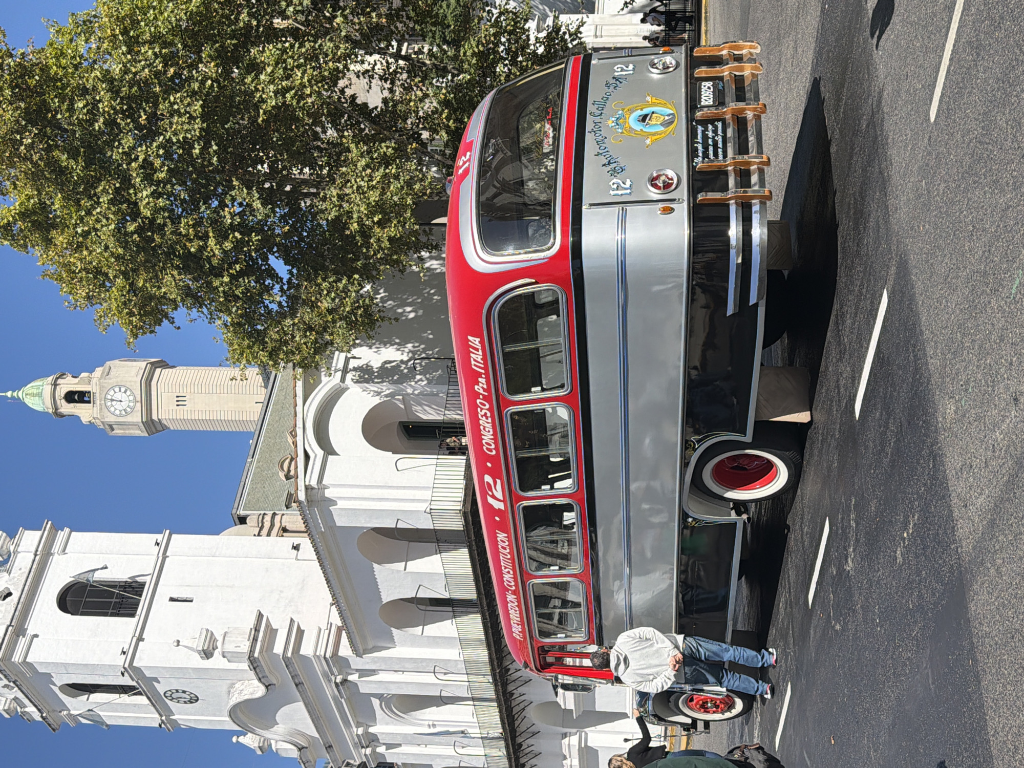 A vintage double-decker bus, predominantly silver with red accents, is parked on a paved street next to a white colonial-style building with arches and a prominent clock tower in the background. The bus, marked with "12" and text including "CONGRESO-Pza. ITALIA," is partially obstructed by two individuals, one bending down near the front of the bus and another standing nearby. The pavement has white lane markings. A large tree with dense foliage overhangs a portion of the scene. The clock tower displays the time as eleven o'clock. The location is likely near the Basilica Nuestra Señora del Pilar in Buenos Aires, Argentina, due to the architectural style of the adjacent building and the presence of a historical tour bus.